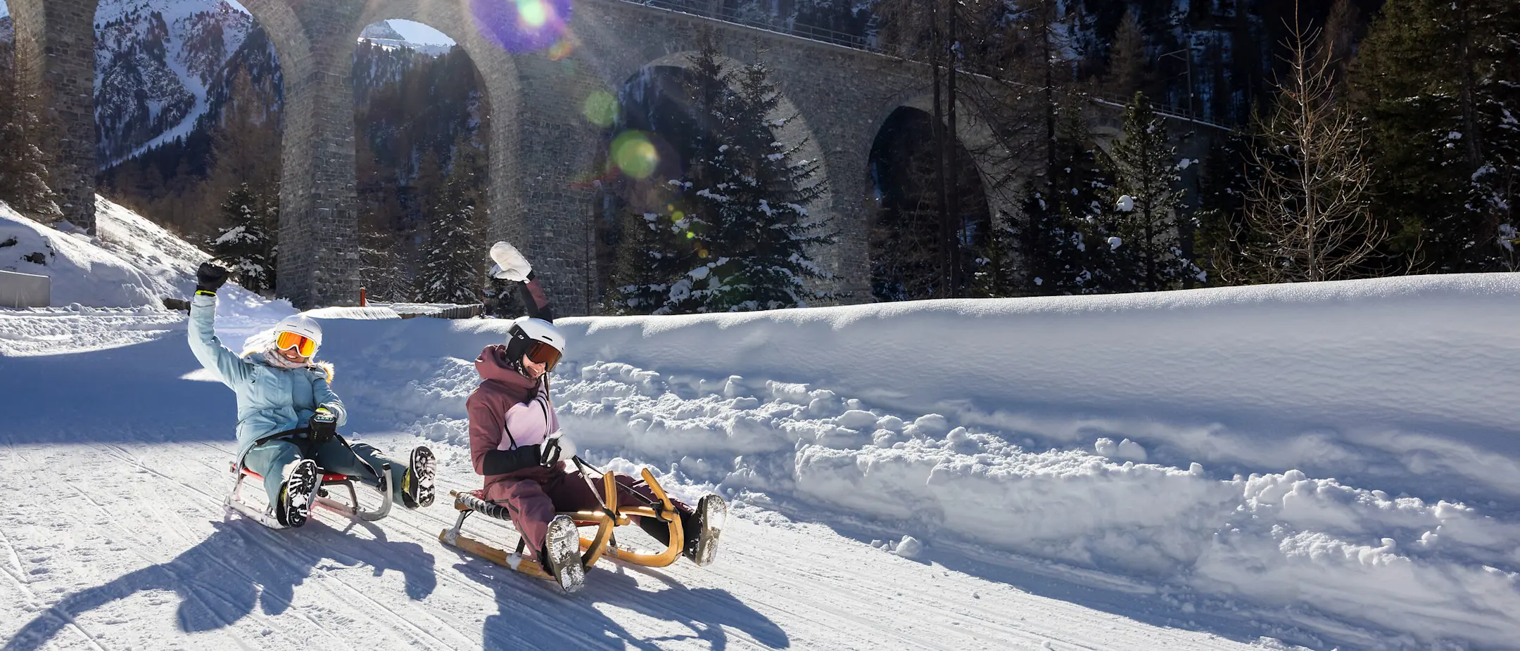 Mehrere Personen rodeln auf einer präparierten Schneepiste unter einem Viadukt hindurch, über den ein roter Zug fährt.