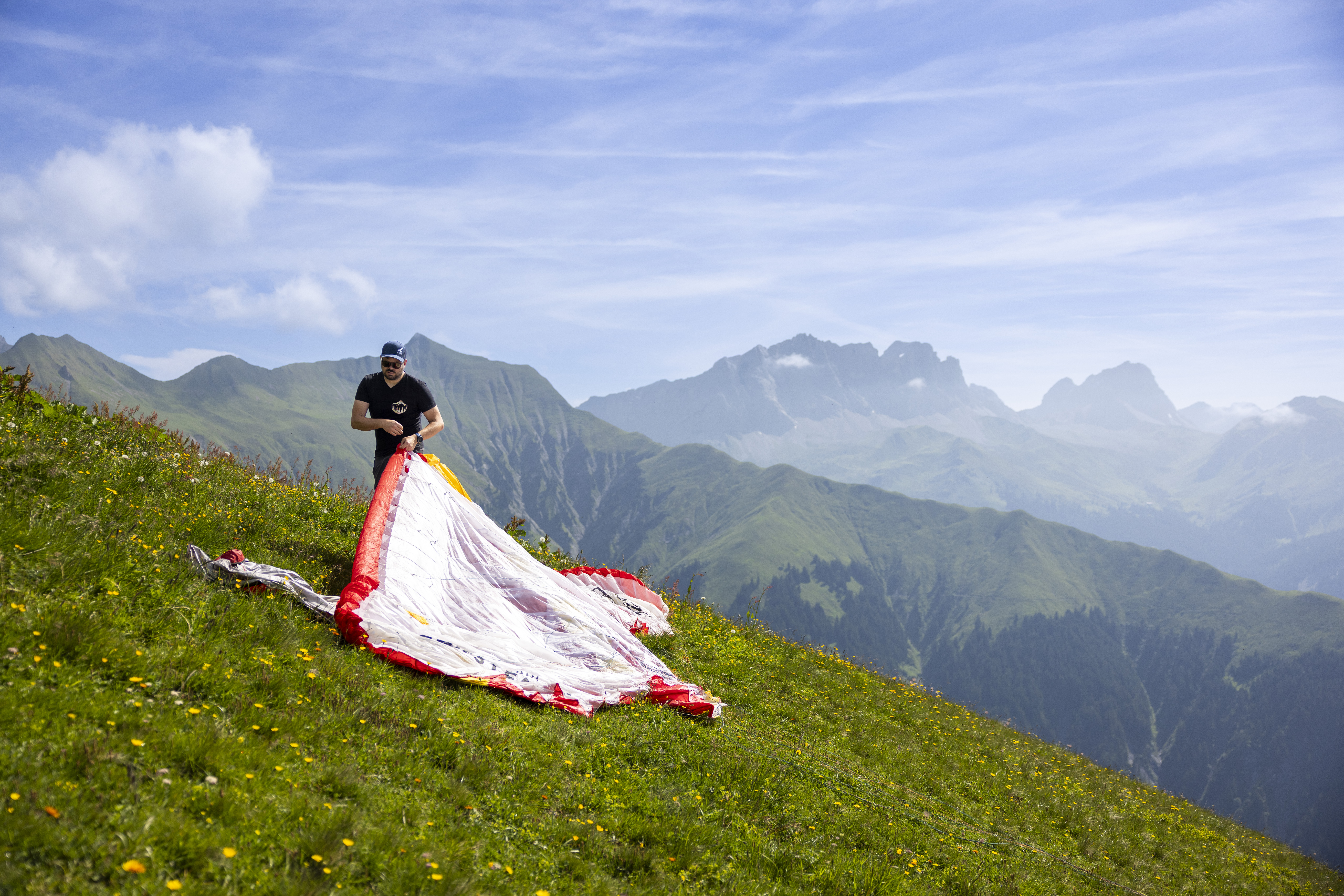 Person bereitet Gleitschirm auf alpiner Wiese vor, mit Blick auf Berglandschaft und weiten Himmel.