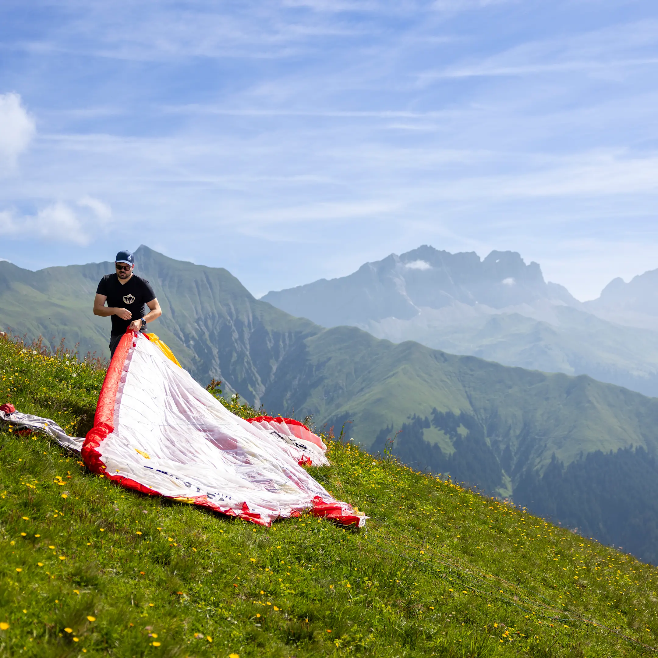 Person bereitet Gleitschirm auf alpiner Wiese vor, mit Blick auf Berglandschaft und weiten Himmel.