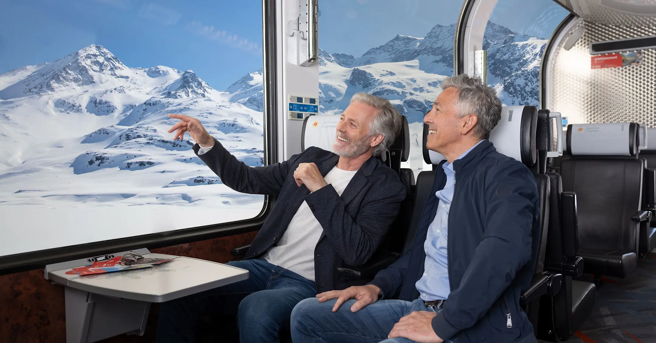 Two men sit in 1st class on the Bernina Express and look out onto the wintry landscape through the panoramic windows.