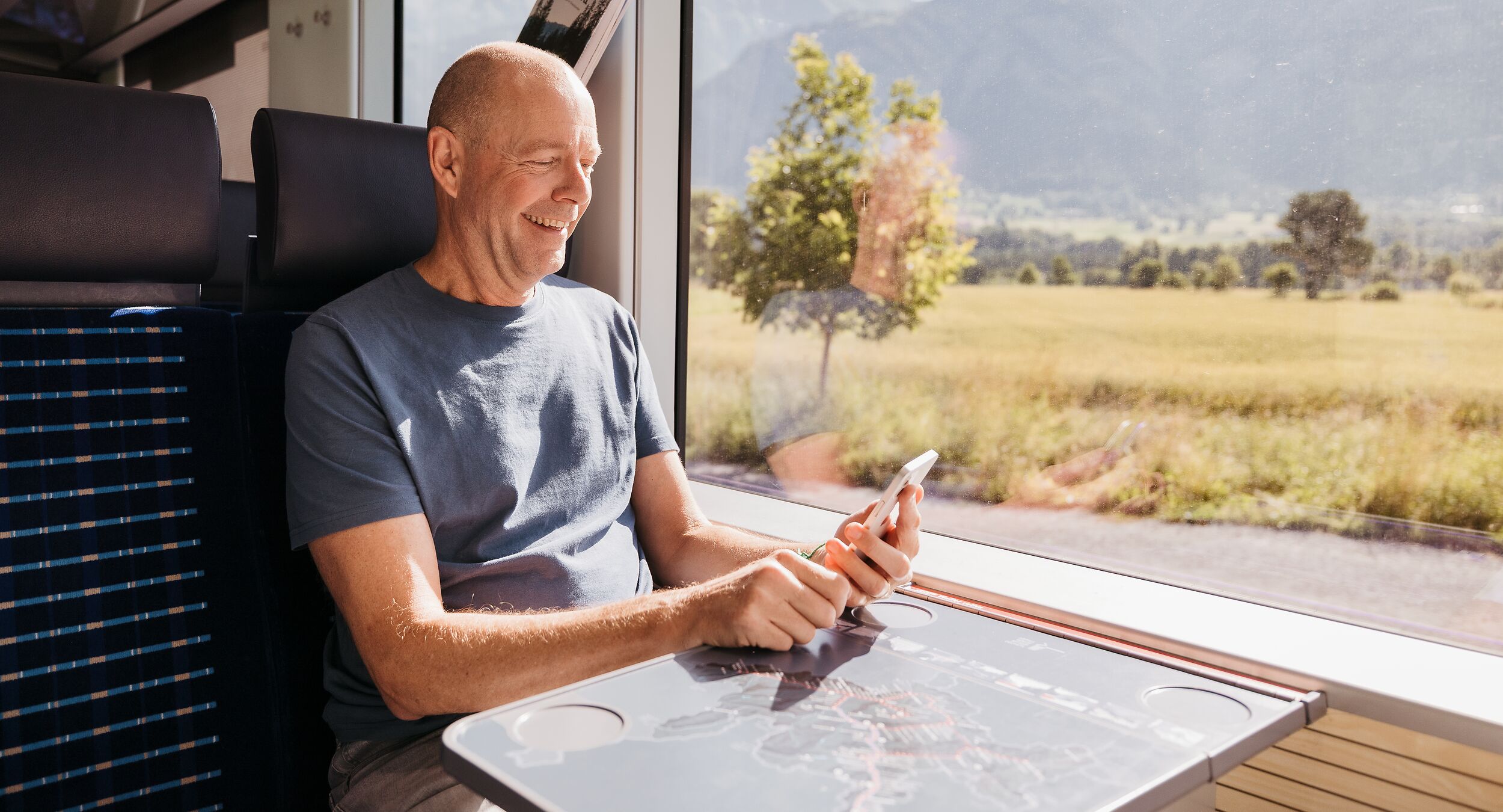 A man is sitting on an RhB regional train and looking at his smartphone with a smile.