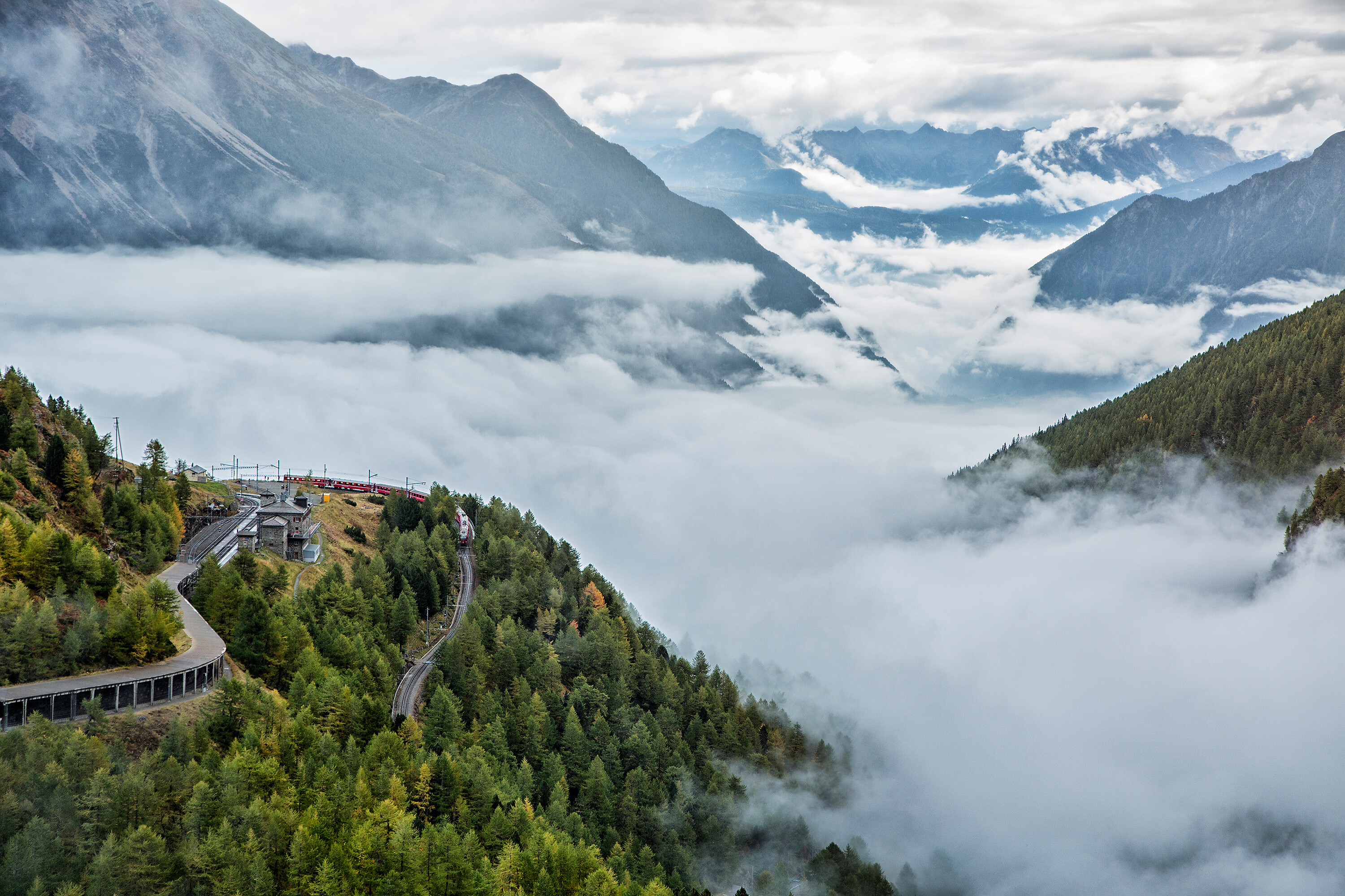 Ein Regionalzug der RhB fährt um die Kurve bei Alp Grüm. Im Hintergrund ist das Valposchiavo von Nebelschwaden bedeckt.