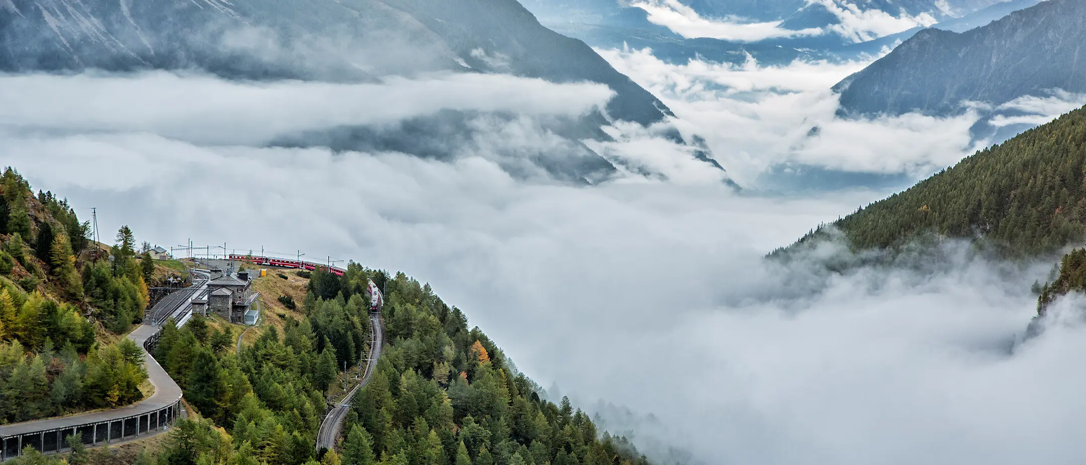 Ein Regionalzug der RhB fährt um die Kurve bei Alp Grüm. Im Hintergrund ist das Valposchiavo von Nebelschwaden bedeckt.