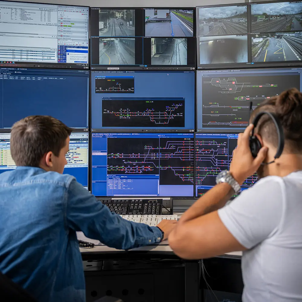 Train dispatcher at work, with a trainee on the left, surrounded by numerous screens