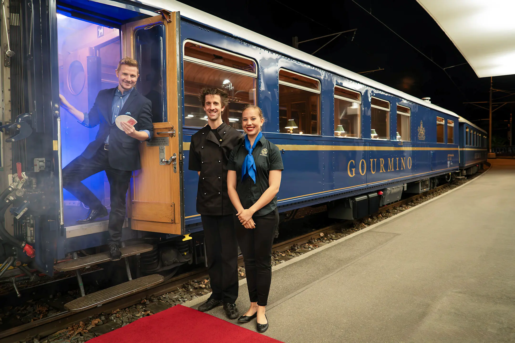 Magier steht mit Spielkarten am blauen Speisewagen Gourmino der RhB. Neben ihm ein Koch und eine Servicekraft.