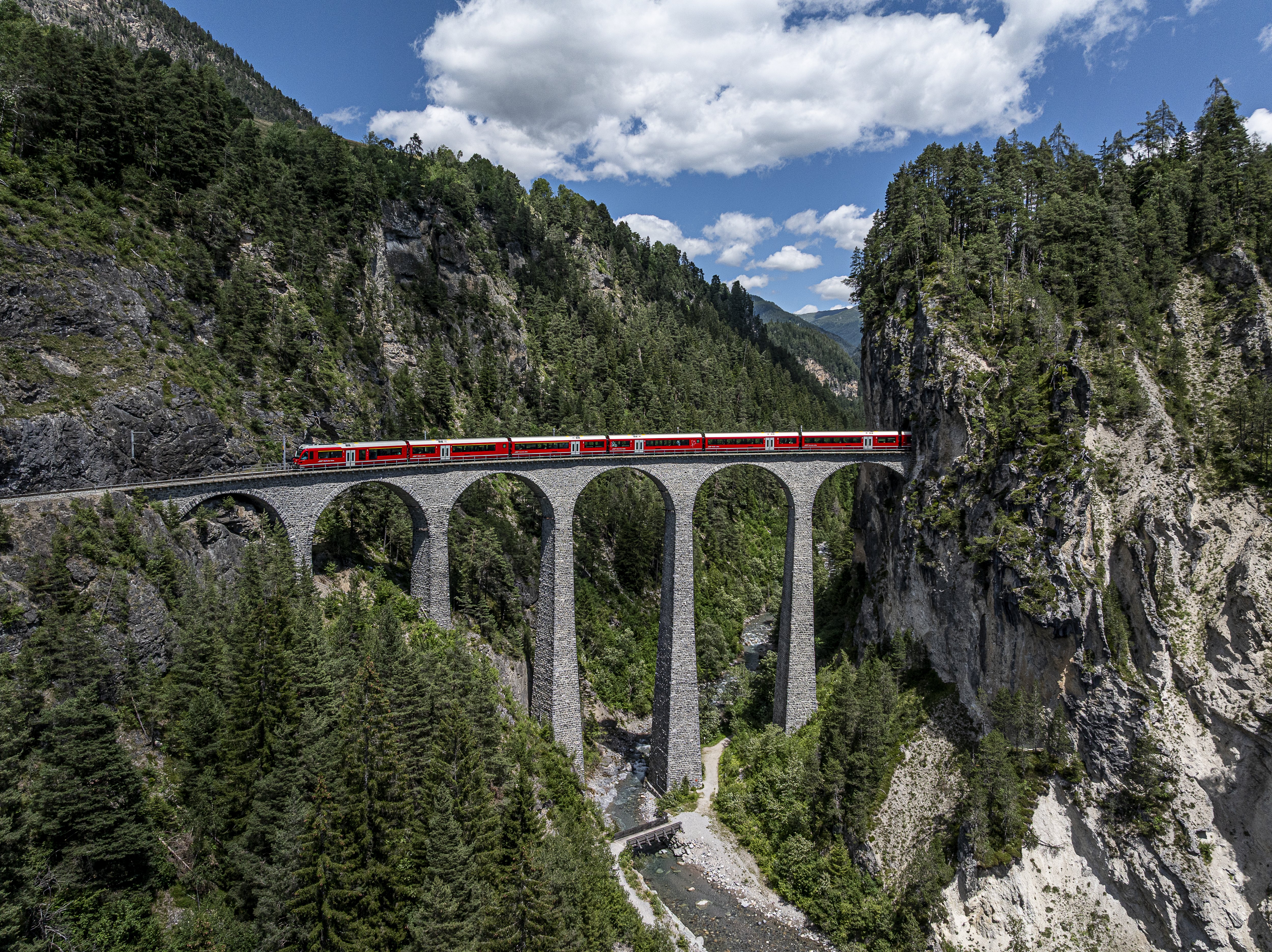 A long red train travels over the RhB Landwasser Viaduct. The bridge spans a deep valley between rock faces.