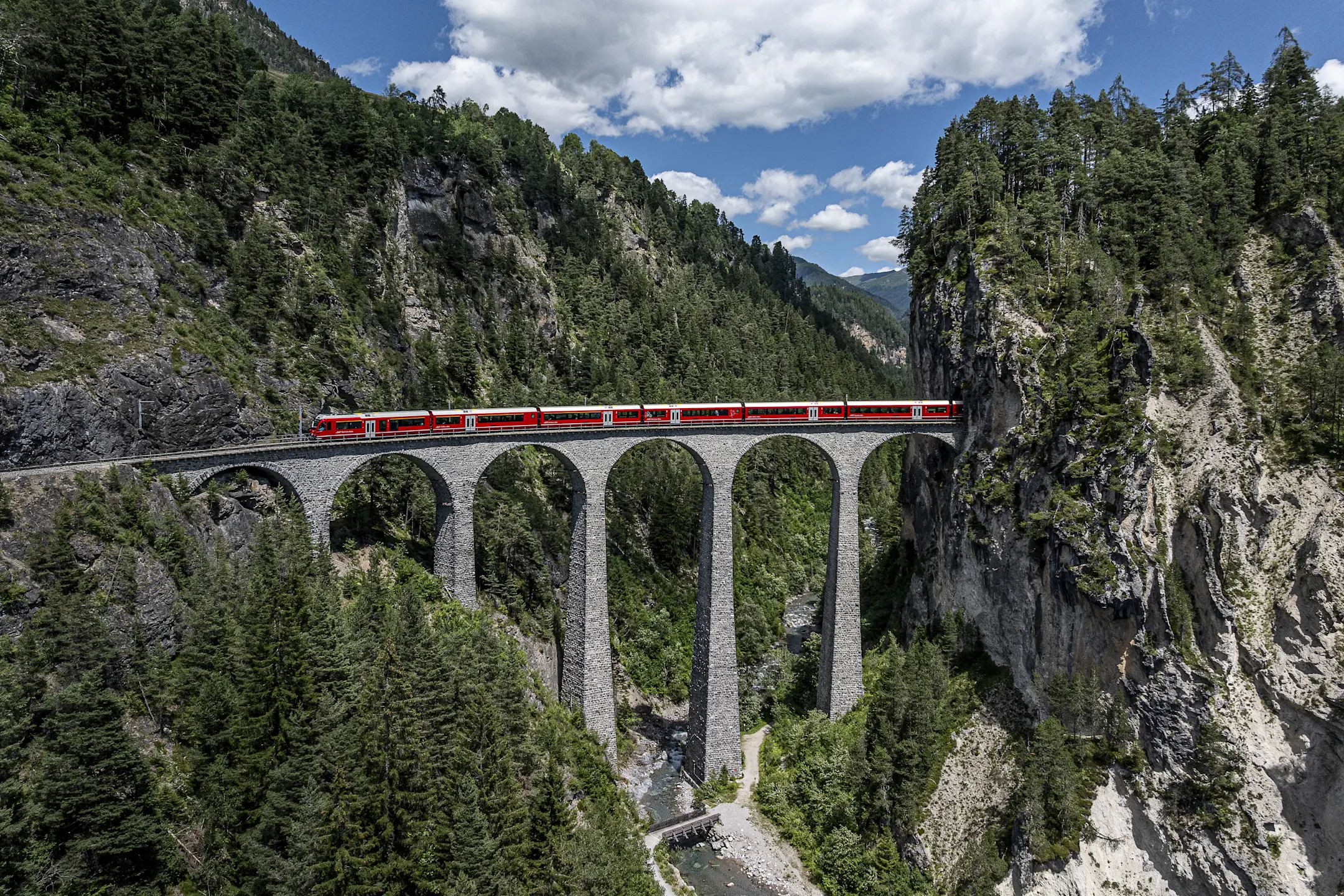 Ein langer roter Zug fährt über das Landwasserviadukt der RhB. Die Brücke überspannt ein tiefes Tal zwischen Felswänden.
