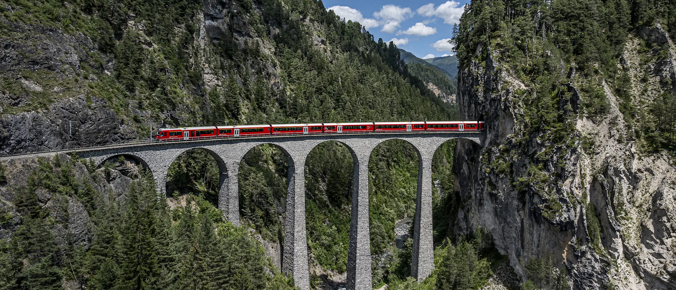 Ein langer roter Zug fährt über das Landwasserviadukt der RhB. Die Brücke überspannt ein tiefes Tal zwischen Felswänden.