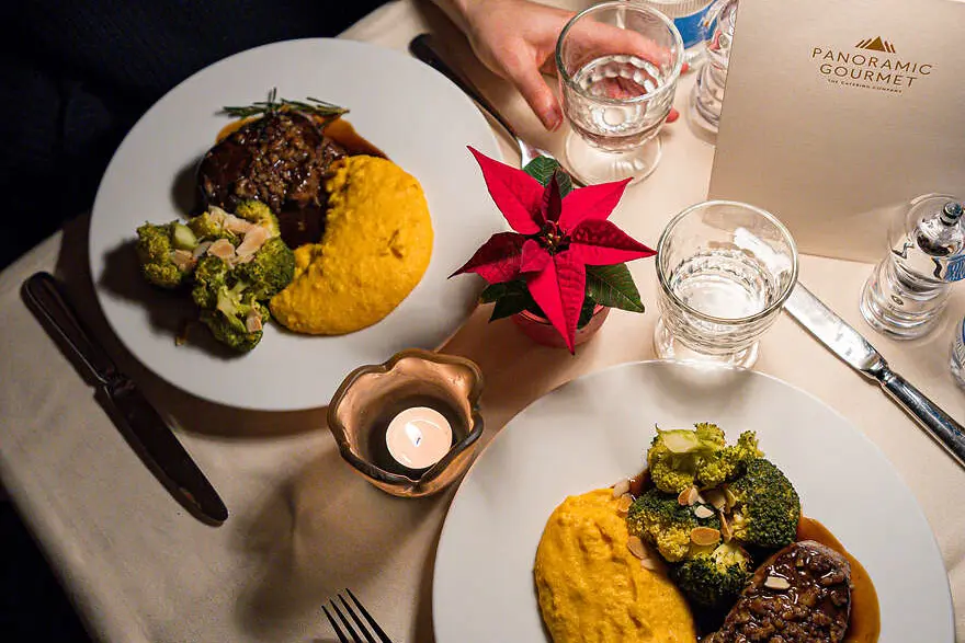 Zwei festlich angerichtete Teller mit Fleisch, Polenta und Gemüse auf einem Tisch im Speisewagen, begleitet von Kerzenlicht.