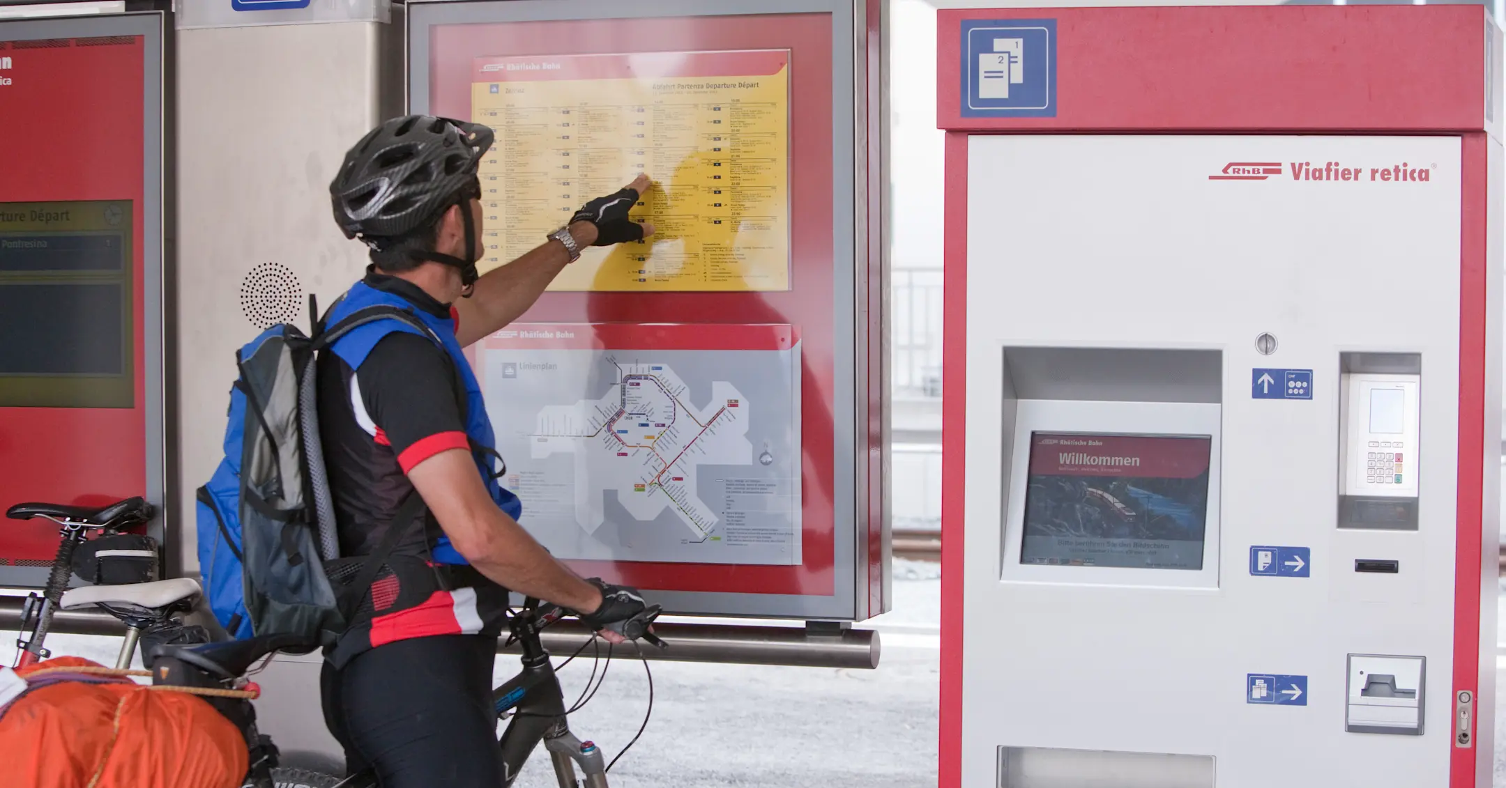 Radfahrer mit Helm und Rucksack informiert sich an einem Fahrplan der Rhätischen Bahn neben einem Ticketautomaten.