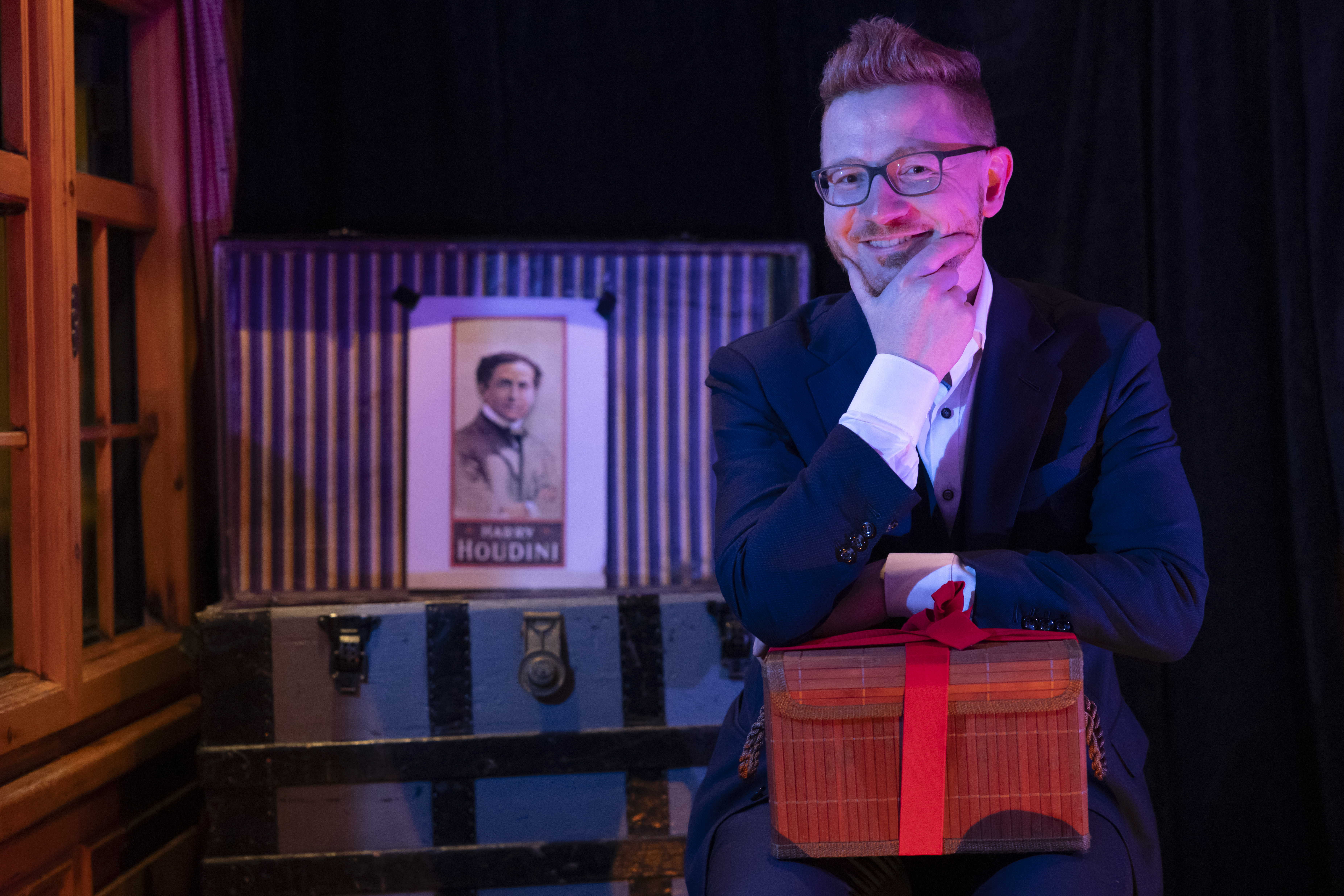 Zauberer Tino Plaz sitzt lächelnd vor einer alten Truhe mit einem Bild von Harry Houdini im Hintergrund. 