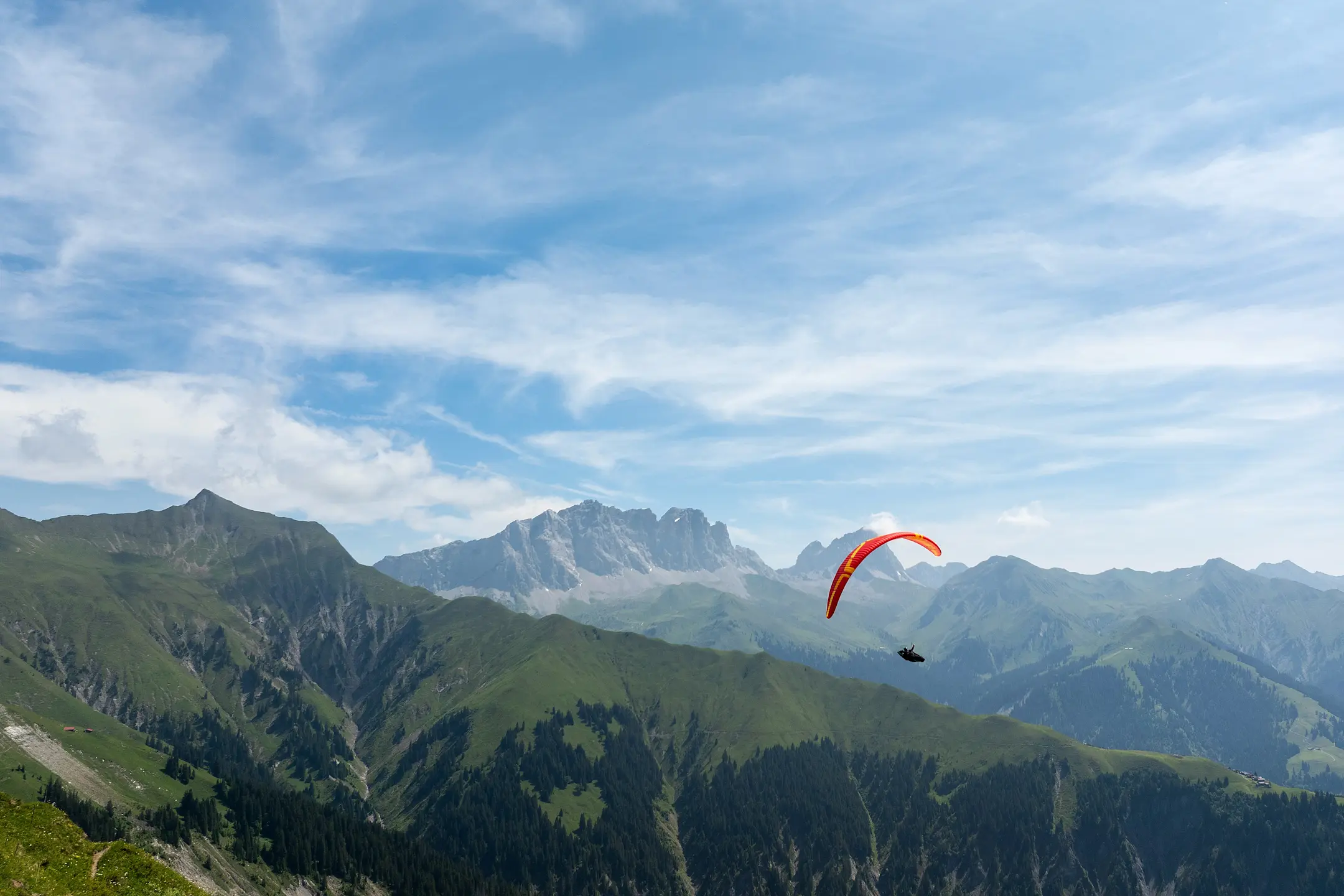 Gleitschirmflieger mit rotem Schirm über grüner Berglandschaft vor alpiner Kulisse und blauem Himmel mit Schleierwolken.