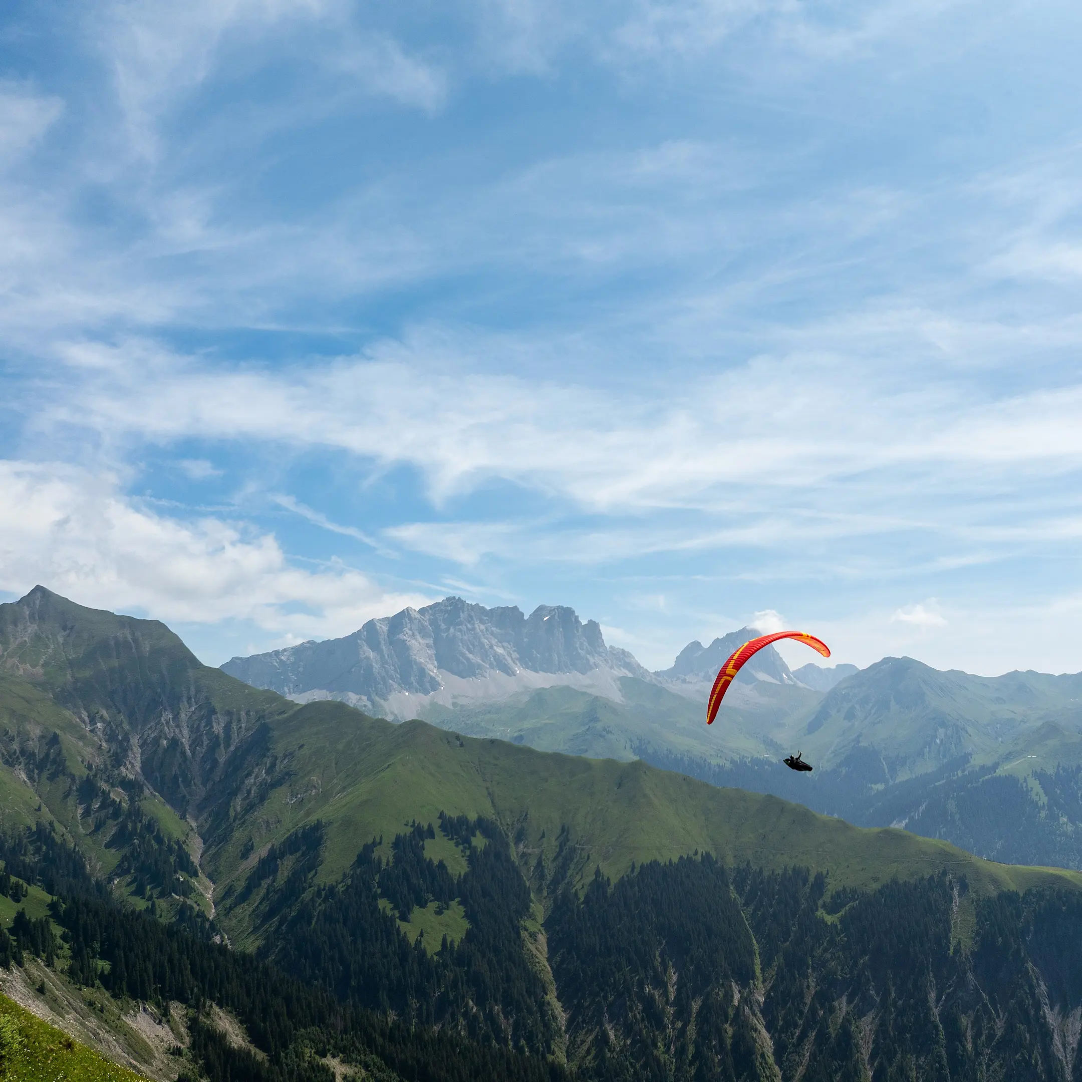 Gleitschirmflieger mit rotem Schirm über grüner Berglandschaft vor alpiner Kulisse und blauem Himmel mit Schleierwolken.