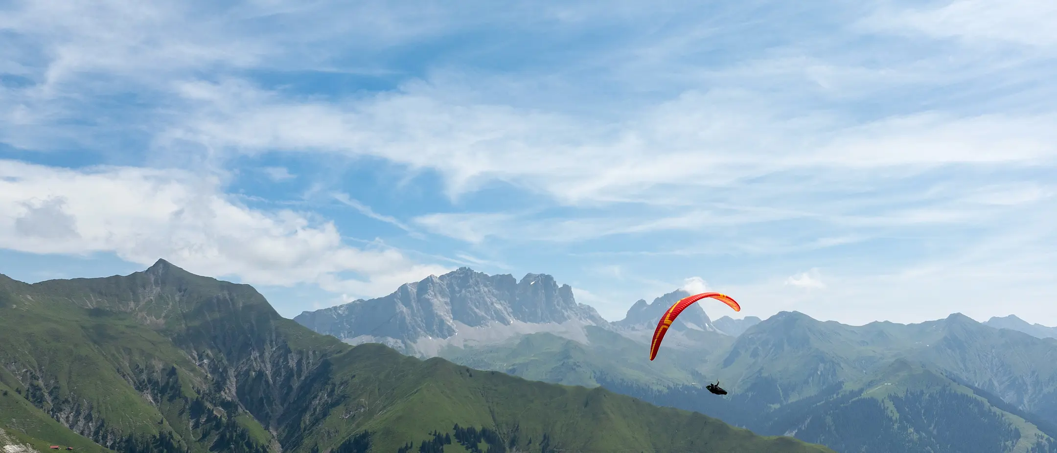 Gleitschirmflieger mit rotem Schirm über grüner Berglandschaft vor alpiner Kulisse und blauem Himmel mit Schleierwolken.