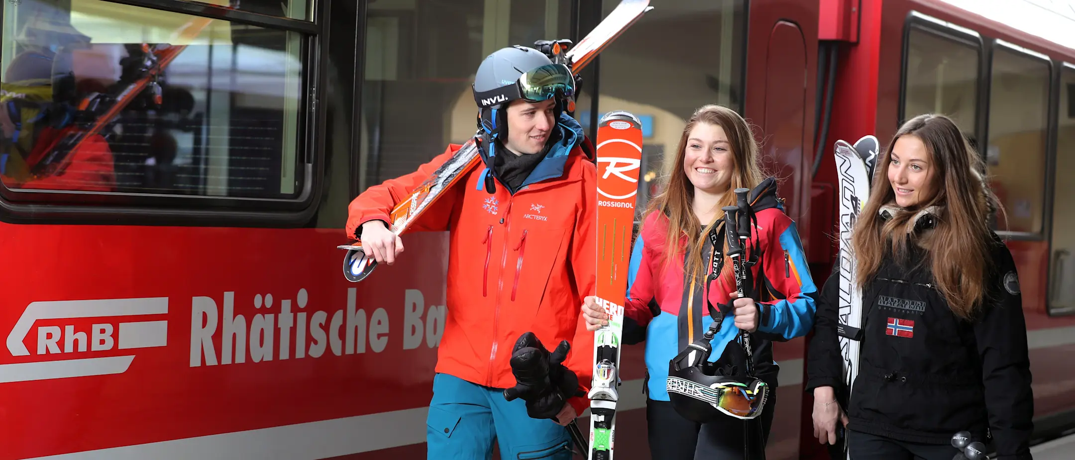 Drei Wintersportler mit Ski- und Snowboardausrüstung steigen lachend aus einem roten Zug der Rhätischen Bahn.