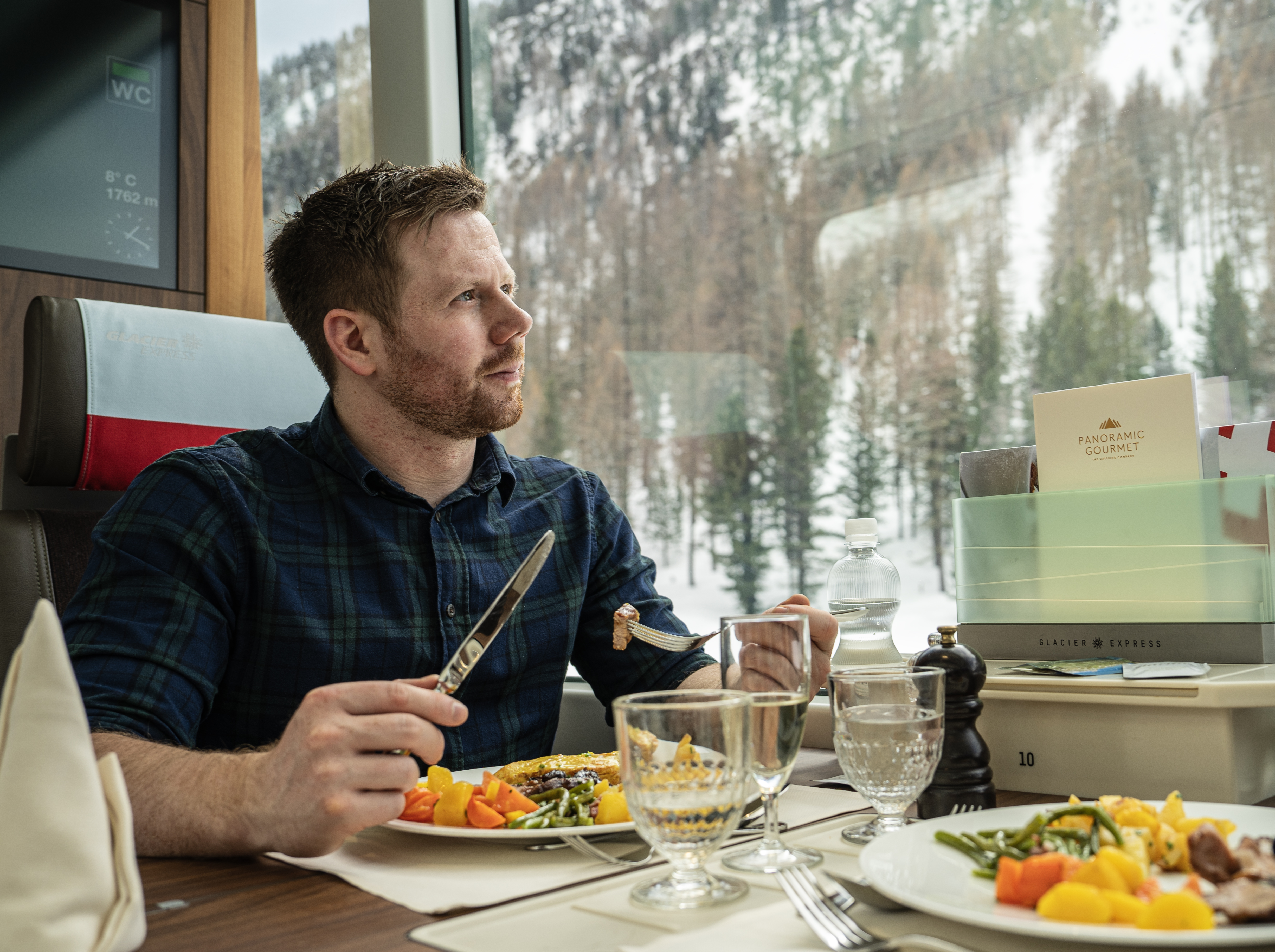Gast geniesst ein mehrgängiges Menü im Panoramawagen des Glacier Express mit Blick auf die verschneite Winterlandschaft.