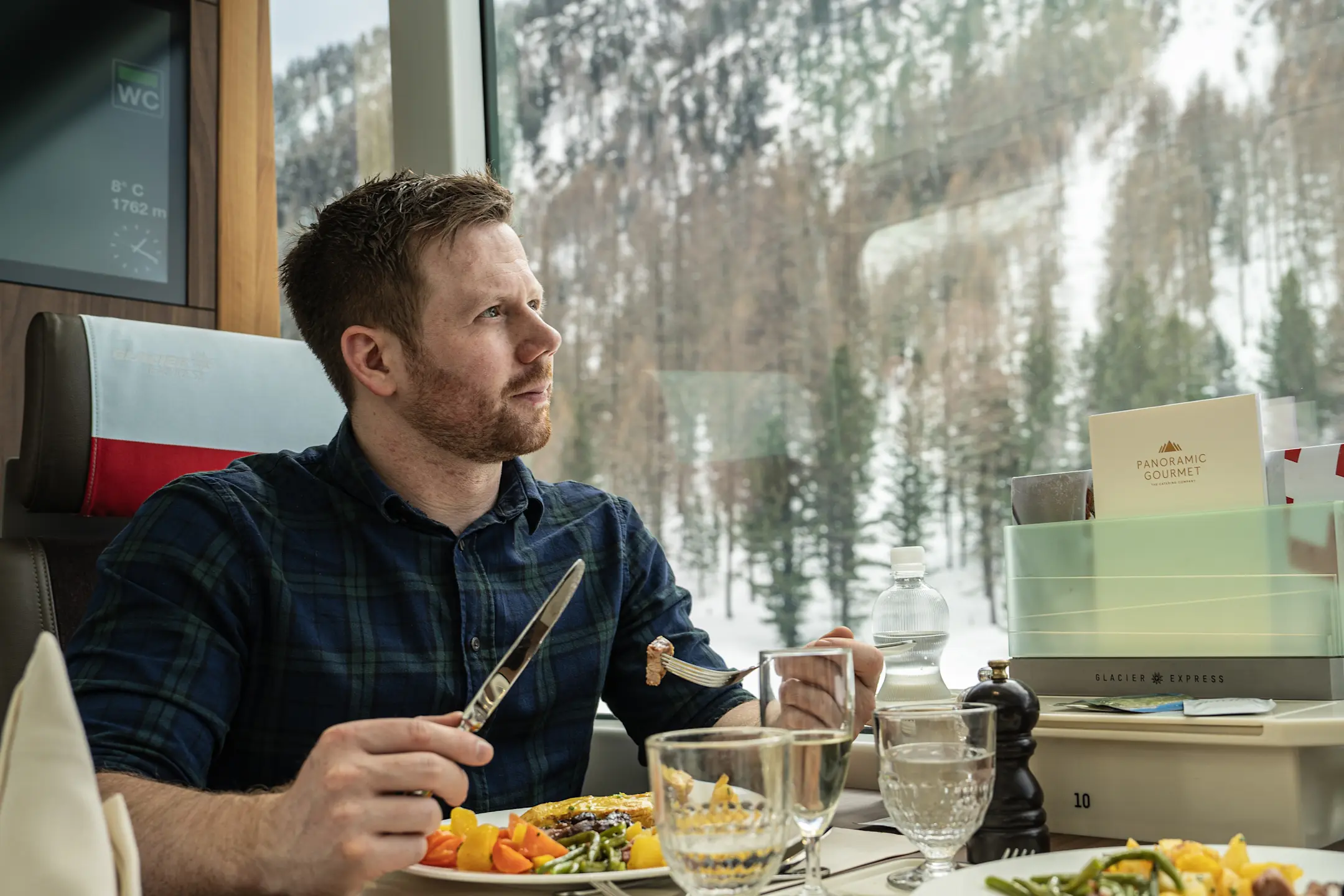 Gast geniesst ein mehrgängiges Menü im Panoramawagen des Glacier Express mit Blick auf die verschneite Winterlandschaft.