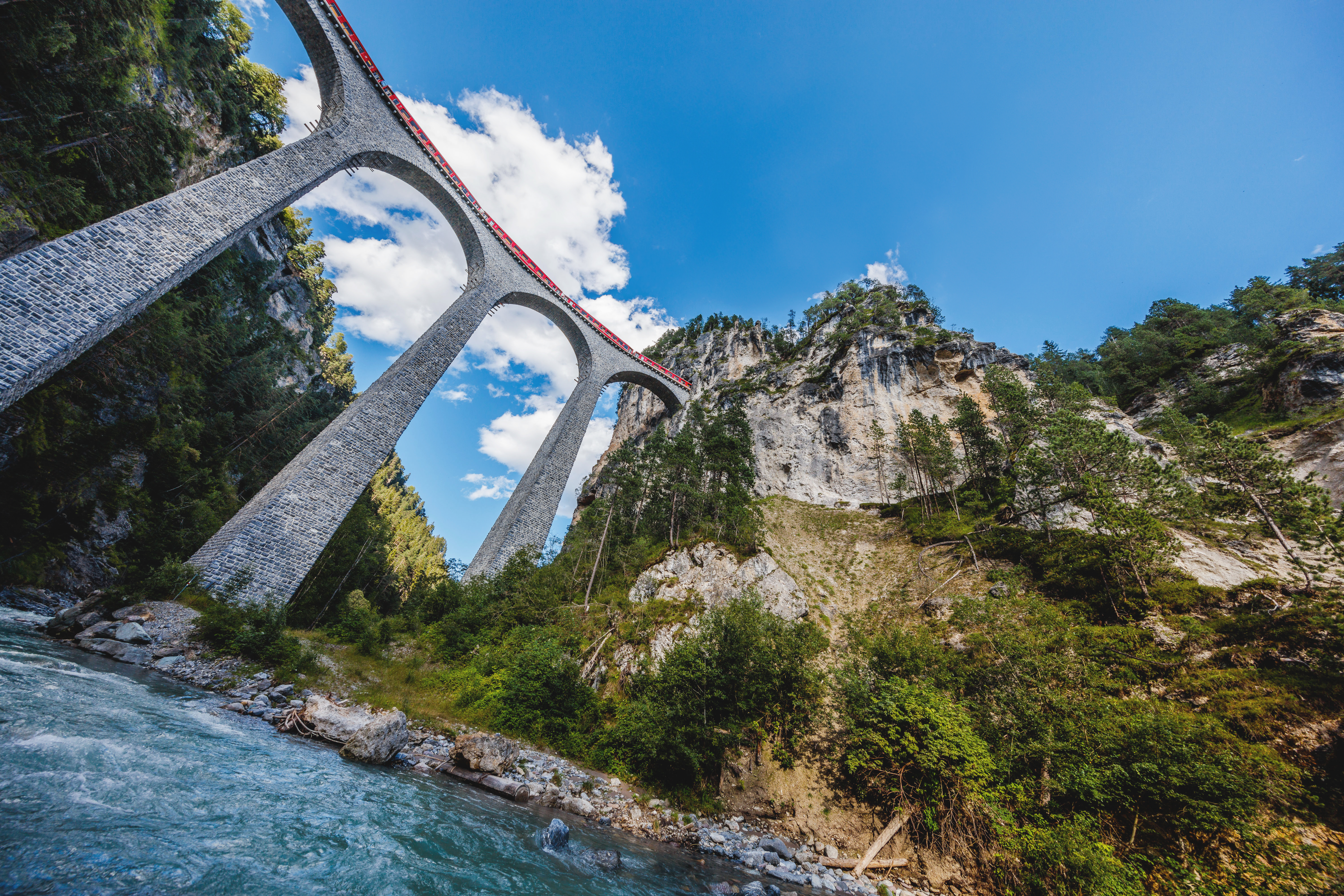 Ein roter Zug überquert das beeindruckende Landwasserviadukt mit hohen Steinbögen, eingebettet in Alpen und Fluss.
