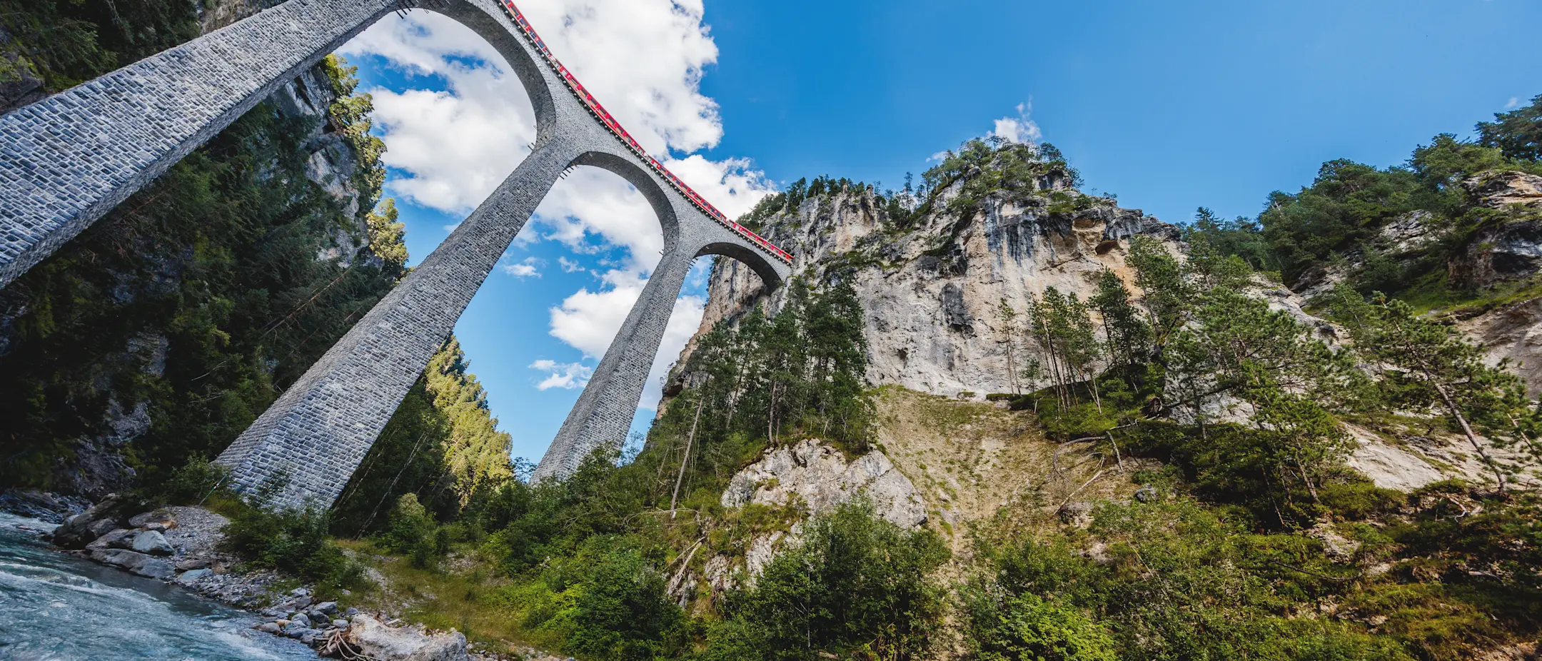 A red train crosses the impressive Landwasser Viaduct with its high stone arches, nestled in the Alps and river.