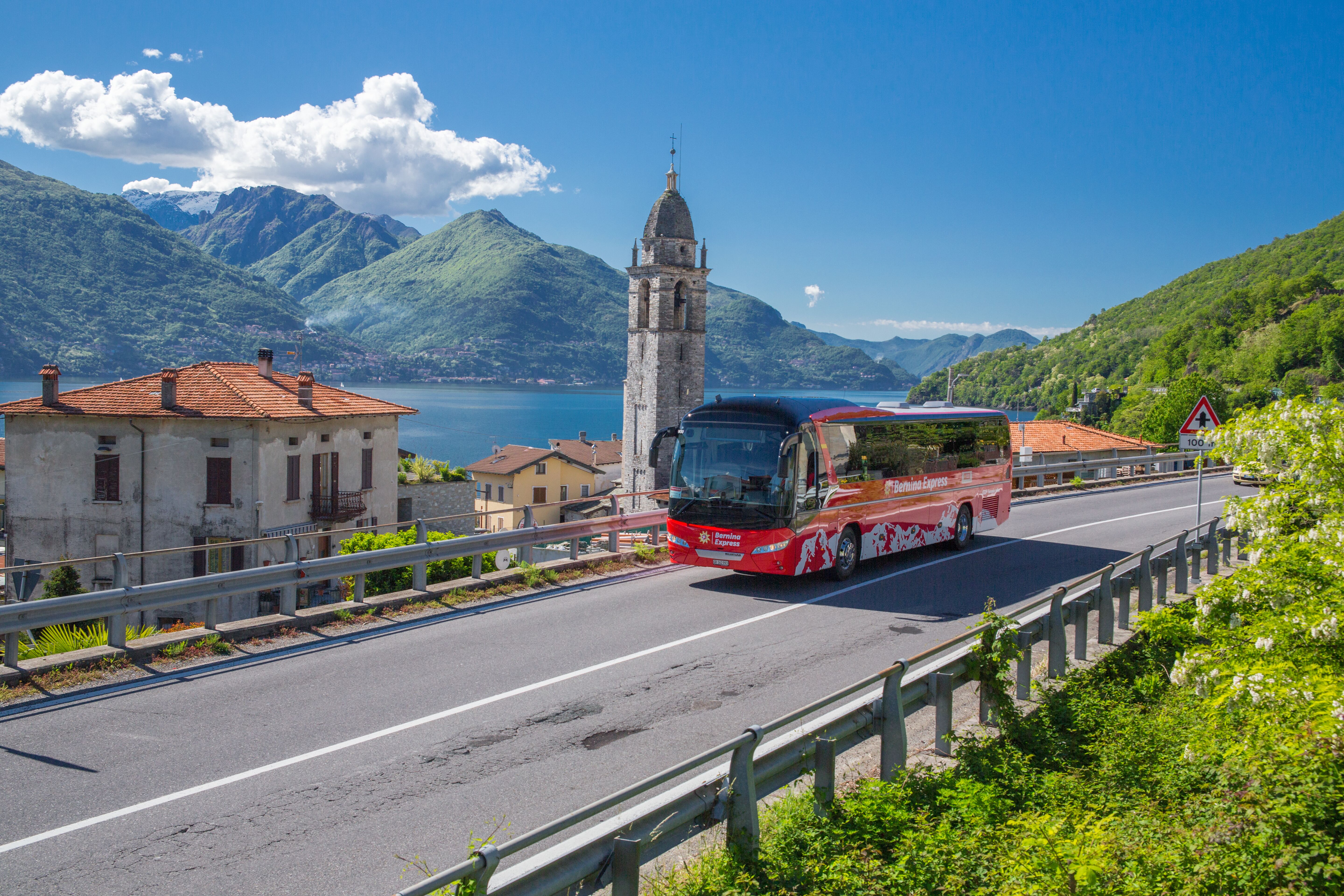 Autobus Bernina Express con tempo soleggiato sul Lago di Como. Pittoresco villaggio con chiesa, lago e montagne sullo sfondo.