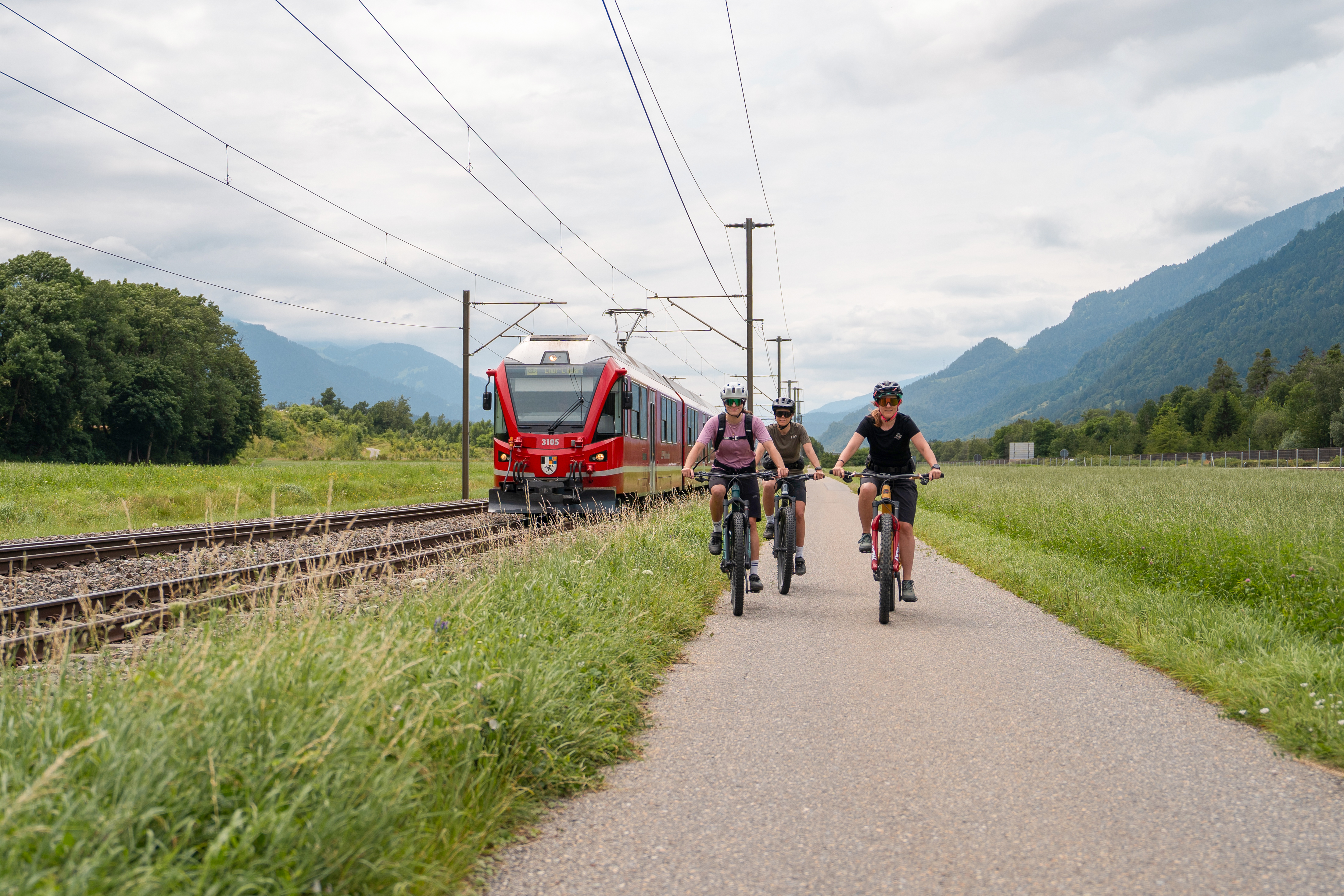 Drei Velofahrer auf einem Biketrail, daneben ein Zug der Rhätischen Bahn.