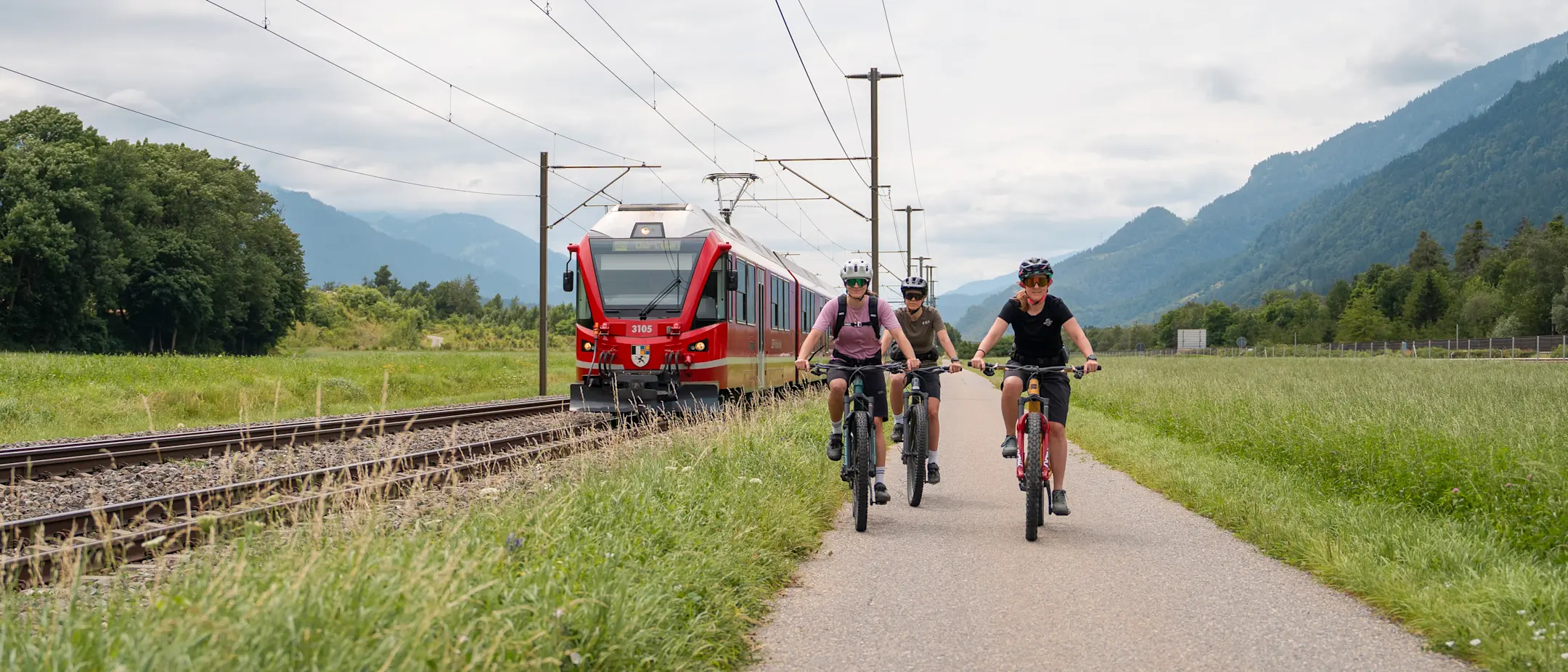 Drei Velofahrer auf einem Biketrail, daneben ein Zug der Rhätischen Bahn.