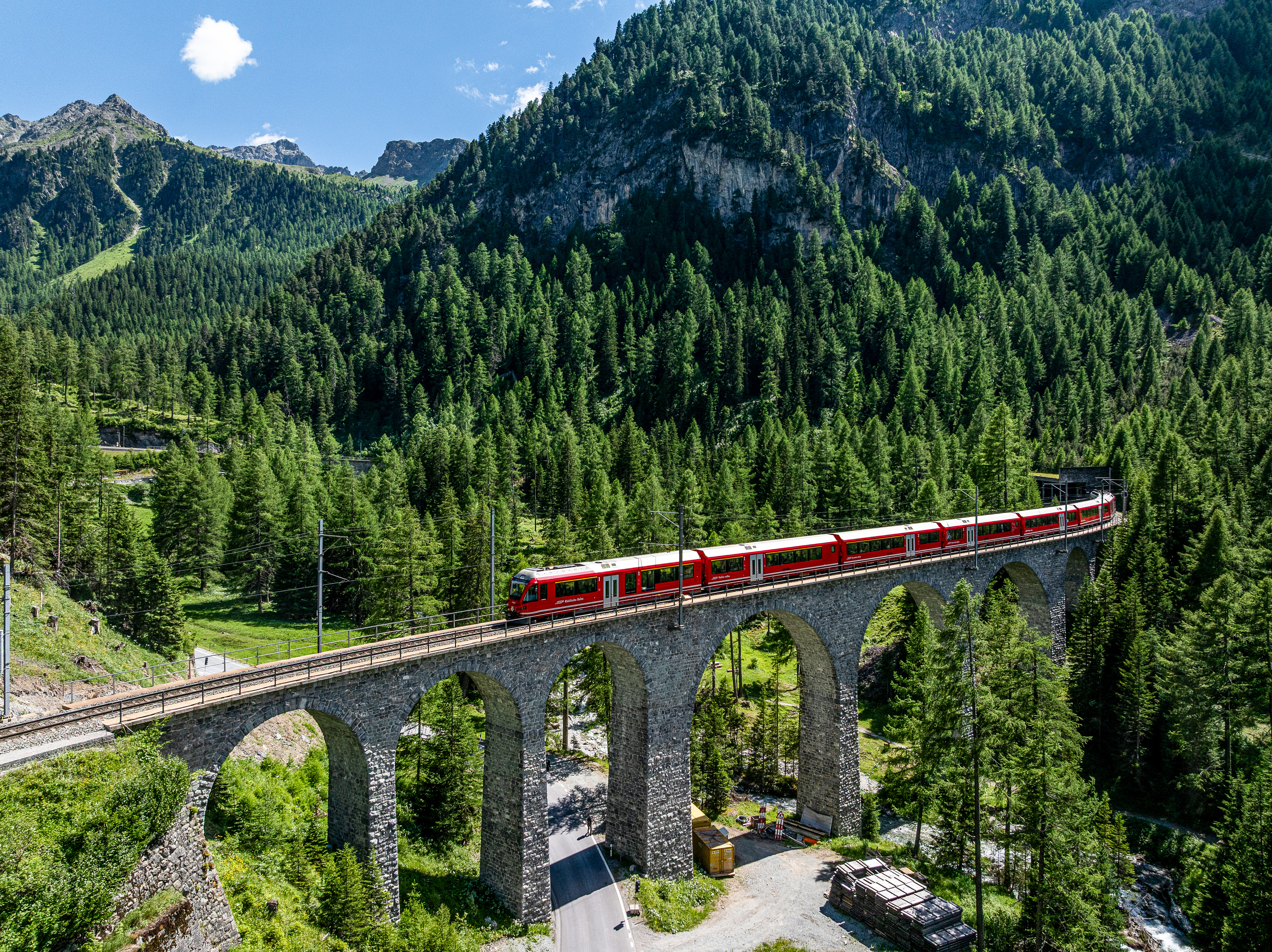 Ein Alvra-Gliederzug der Rhätischen Bahn überquert ein Viadukt im grünen Albulatal, eingebettet in die Bergwelt Graubündens.