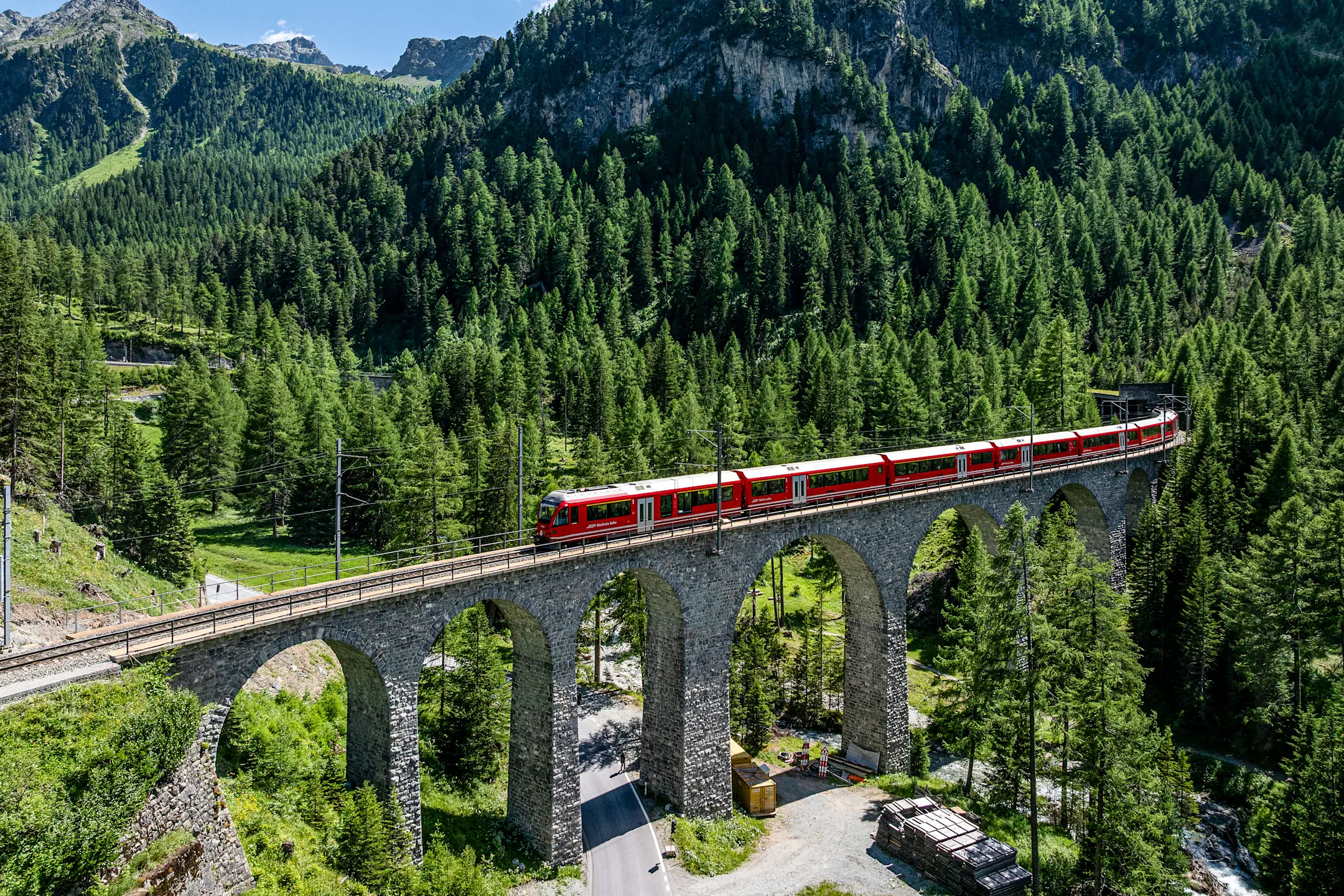 Ein Alvra-Gliederzug der Rhätischen Bahn überquert ein Viadukt im grünen Albulatal, eingebettet in die Bergwelt Graubündens.