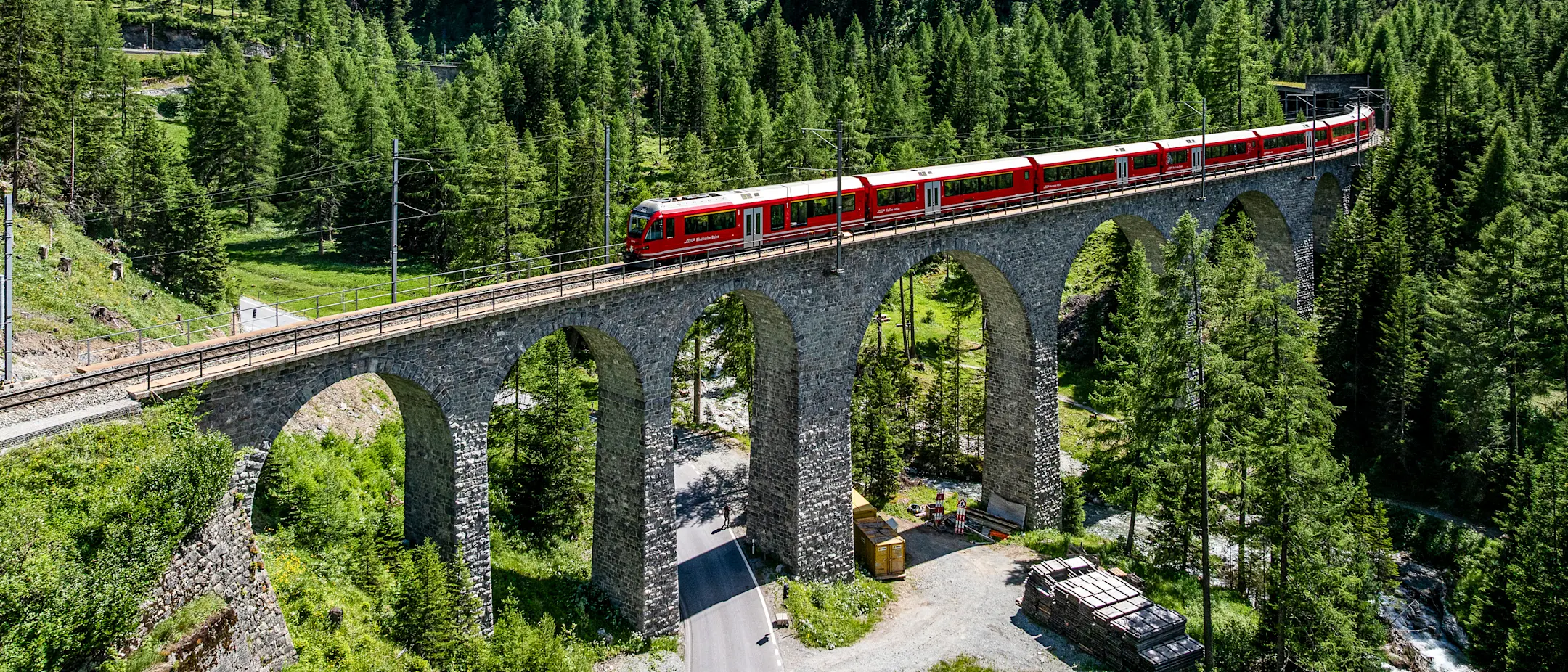 Ein Alvra-Gliederzug der Rhätischen Bahn überquert ein Viadukt im grünen Albulatal, eingebettet in die Bergwelt Graubündens.