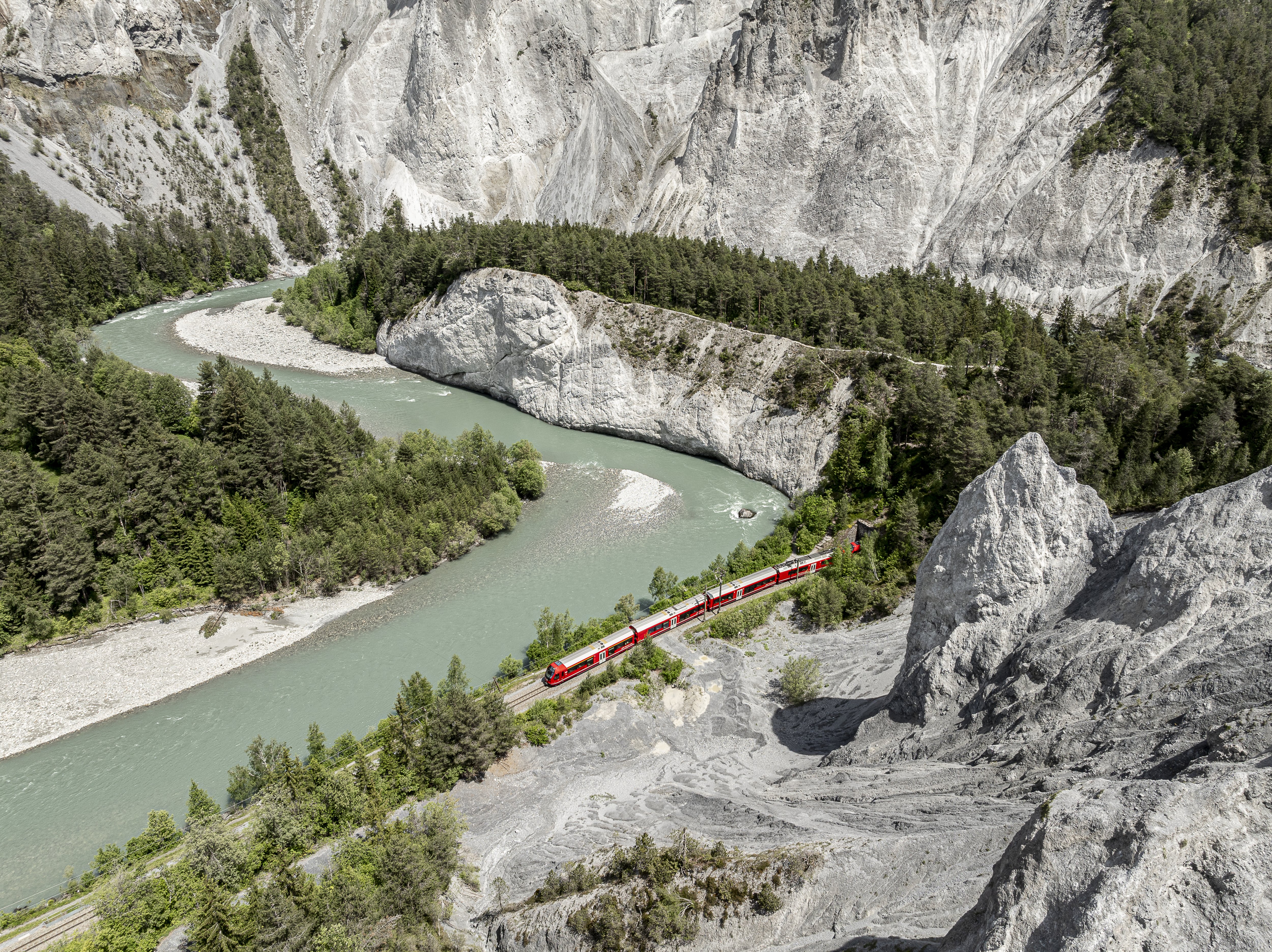 Ein Zug der Rhätischen Bahn fährt entlang des türkisgrünen Rheins durch die Rheinschlucht mit hellen Felswänden.
