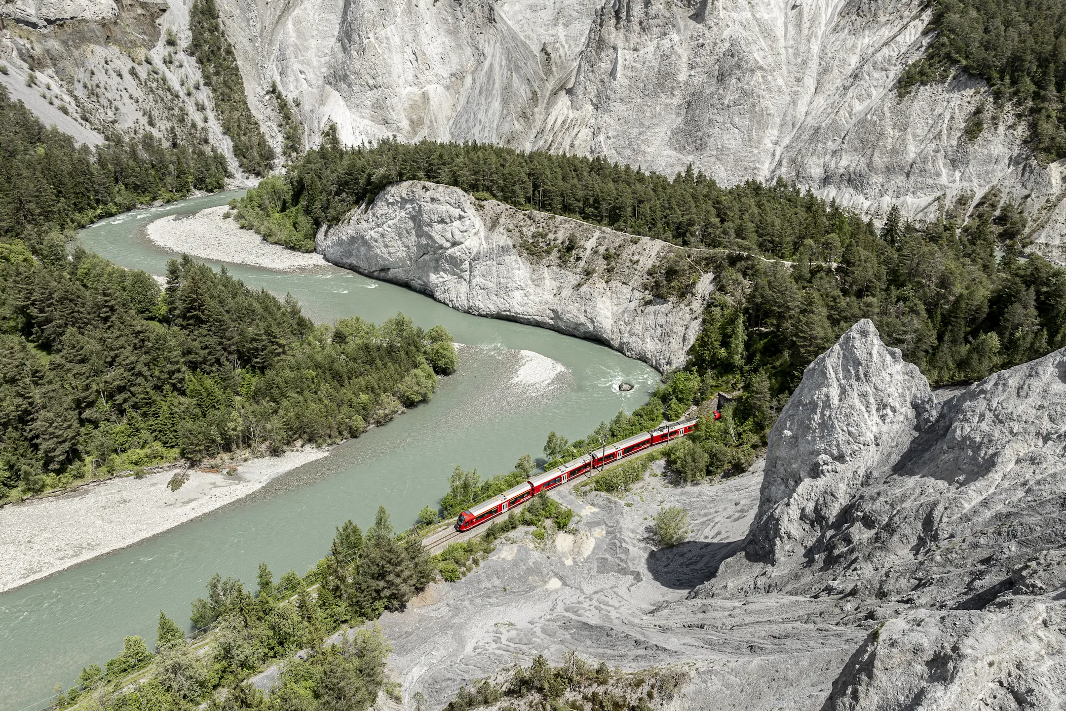 Ein Zug der Rhätischen Bahn fährt entlang des türkisgrünen Rheins durch die Rheinschlucht mit hellen Felswänden.