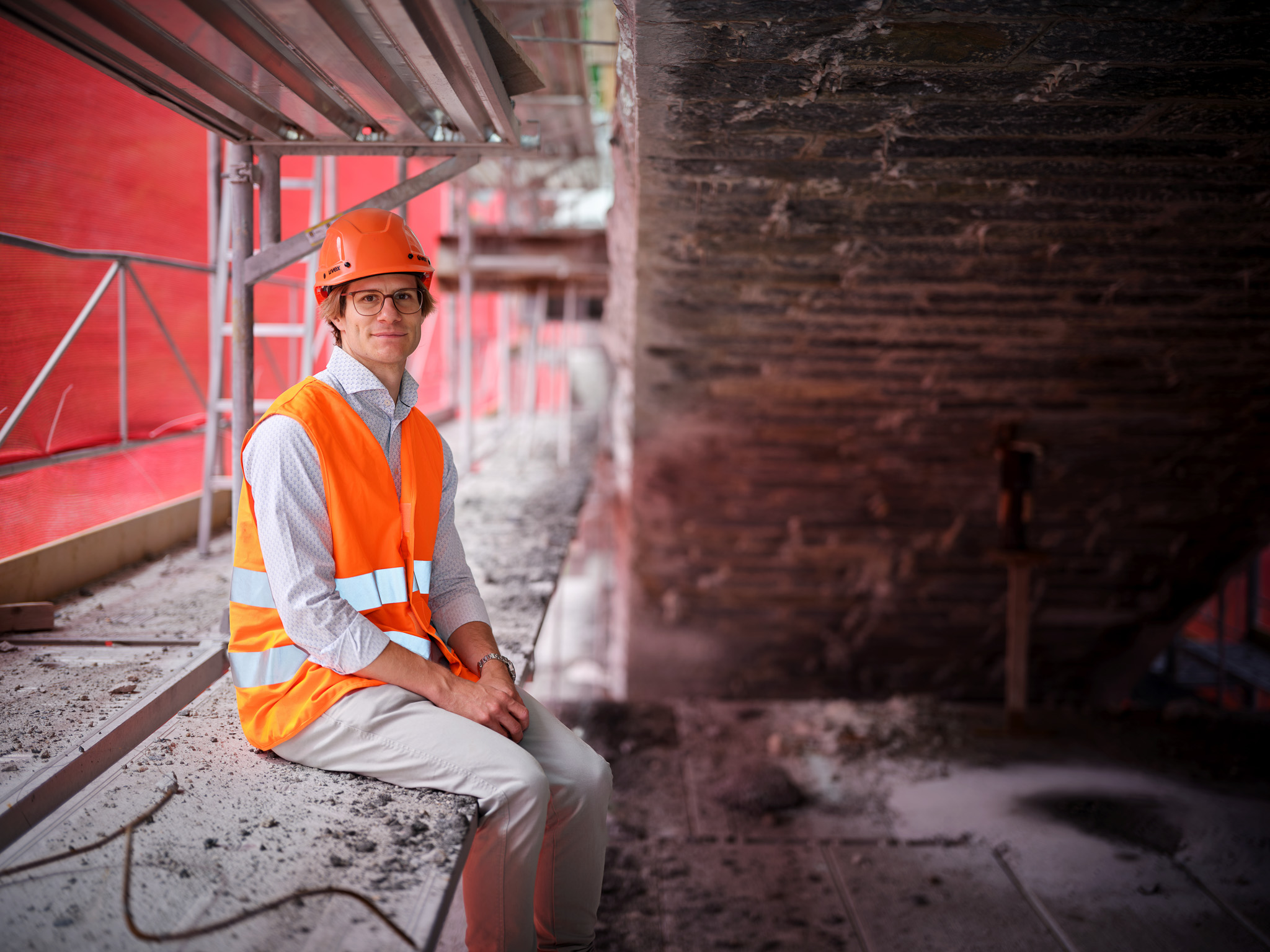 Person in orangefarbener Schutzweste und Helm sitzt auf Baustellengerüst unter einem gemauerten Tunnelgewölbe.