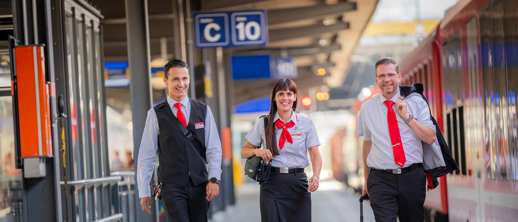 Drei lächelnde RhB-Zugbegleiter:innen laufen über einen Bahnsteig. Eine Frau in der Mitte, zwei Männer außen.
