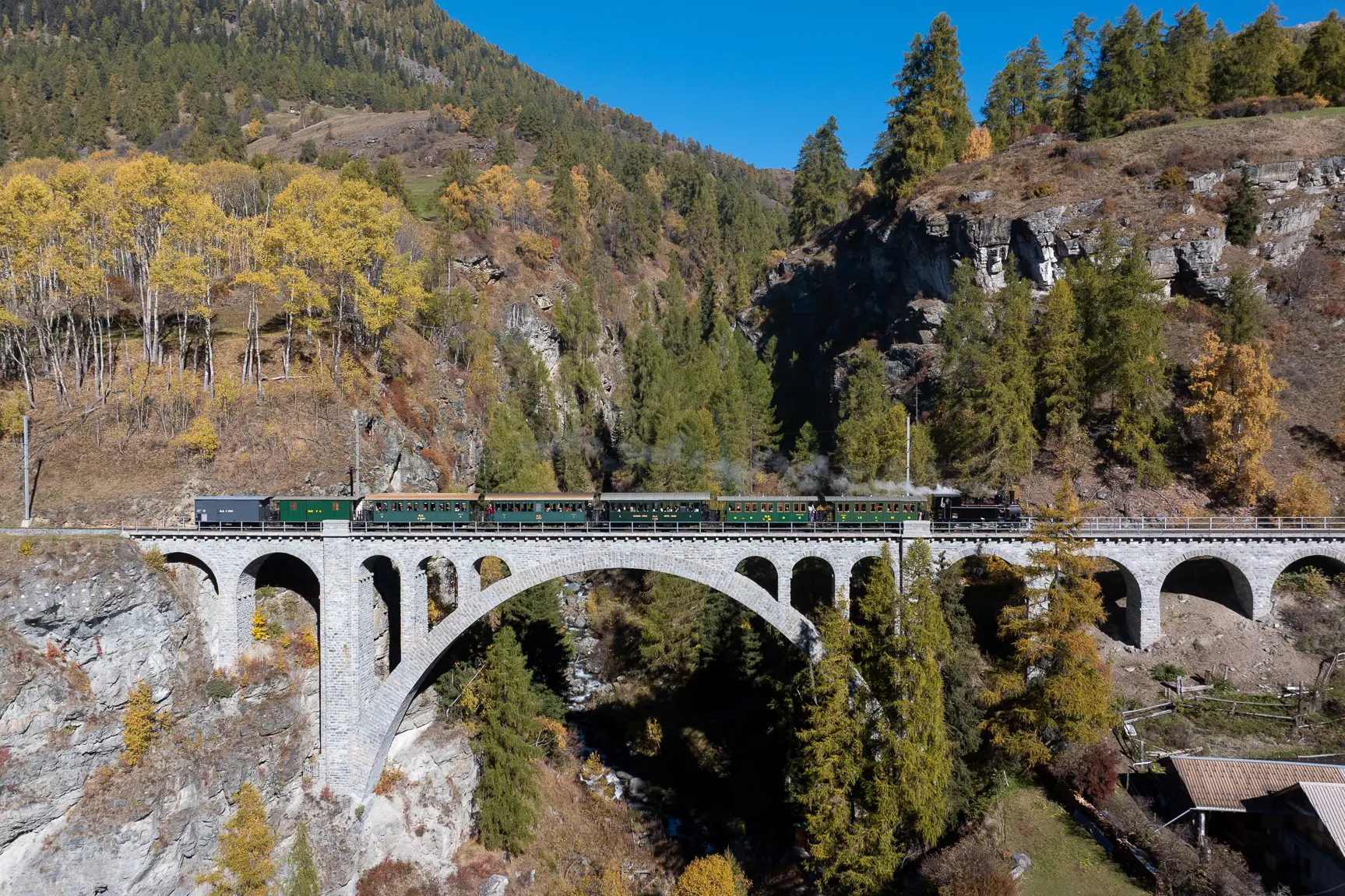 Die historische Dampflok Heidi überquert das Val Tuoi Viadukt vor herbstlicher Bergkulisse.