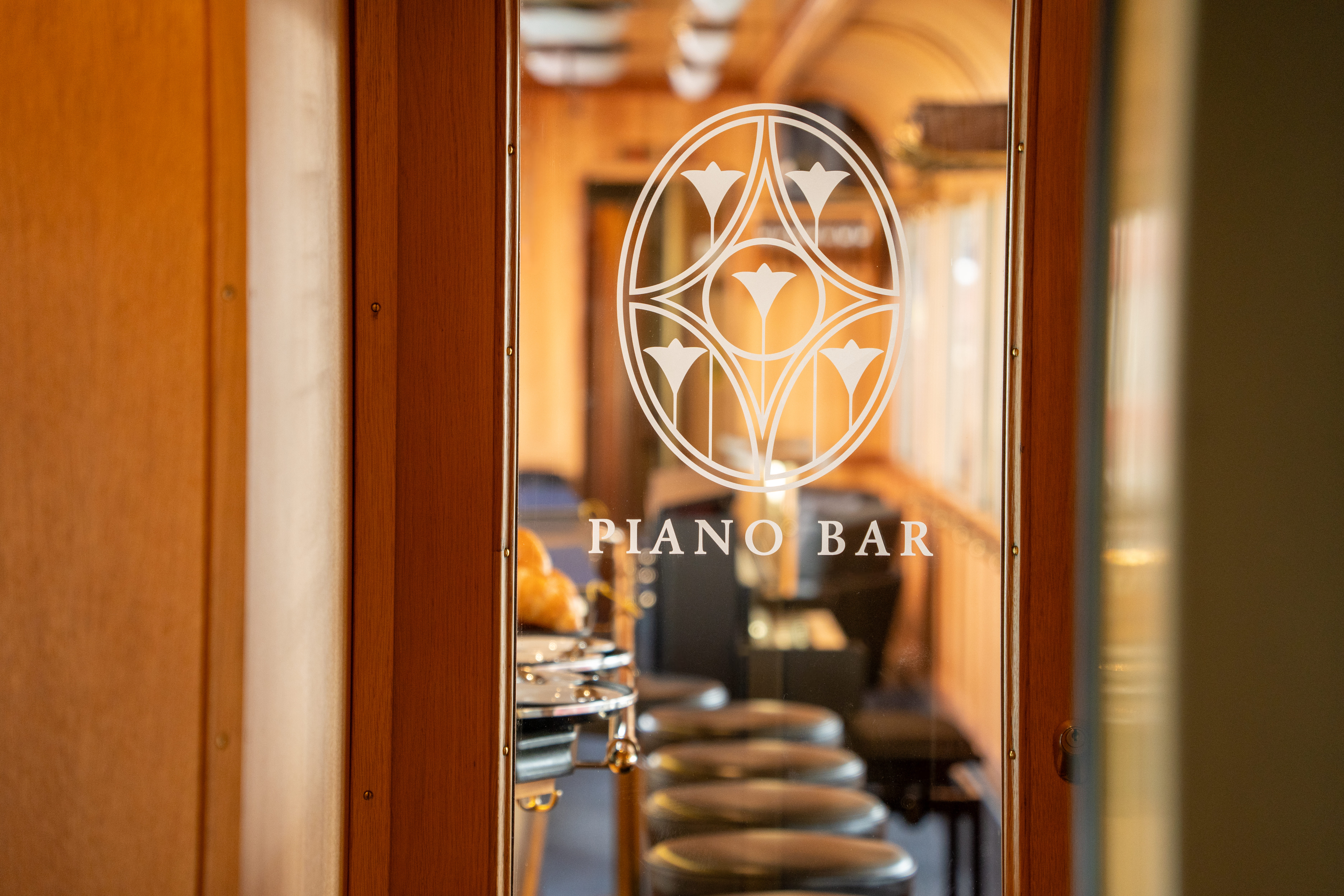 Blick durch eine Glastür mit der Aufschrift „Piano Bar“ in einen stilvollen Zugwaggon mit Barhockern und Holzinterieur.