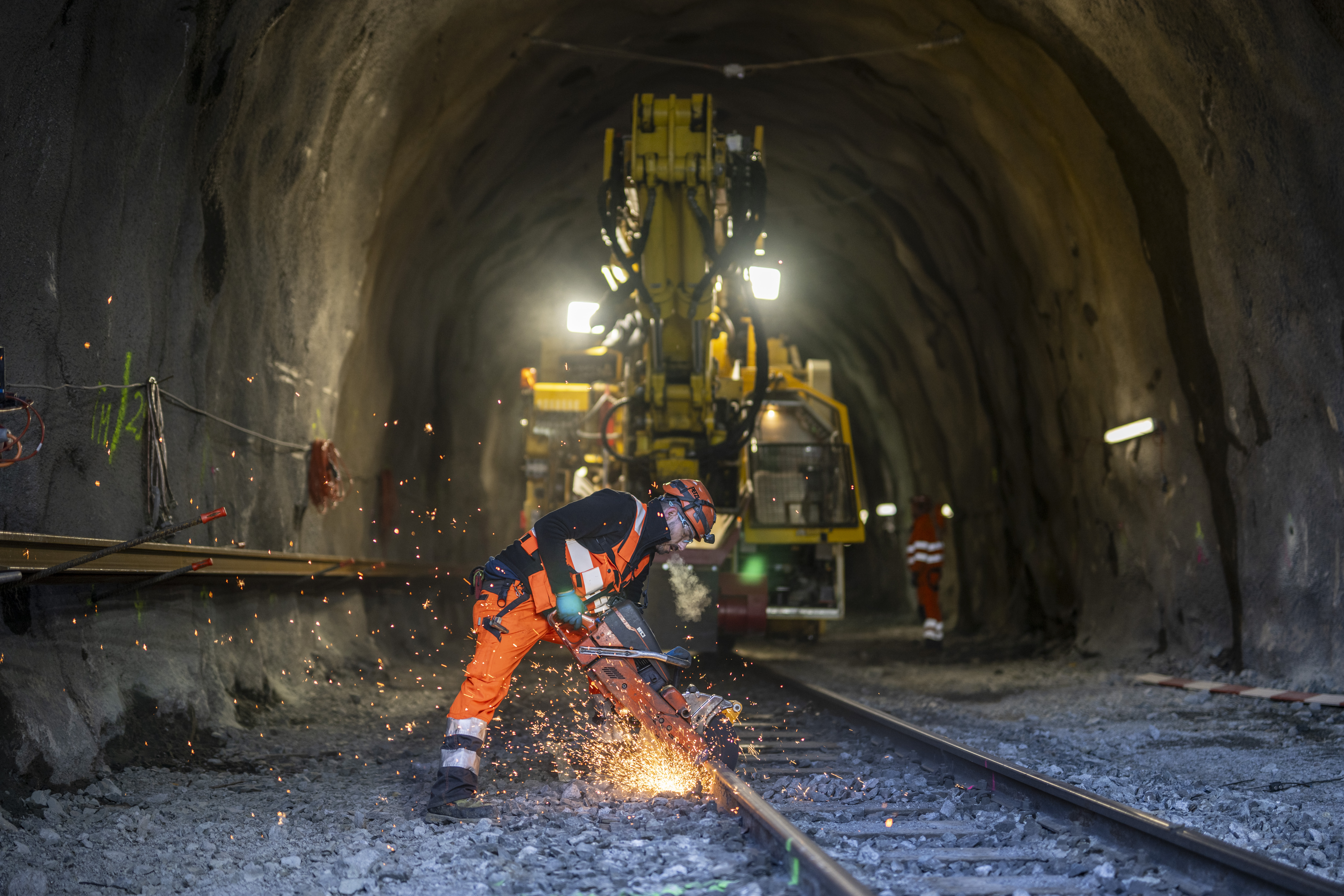 Workers in protective clothing cut tracks in a tunnel with a cut-off grinder. In the background, a construction machine.