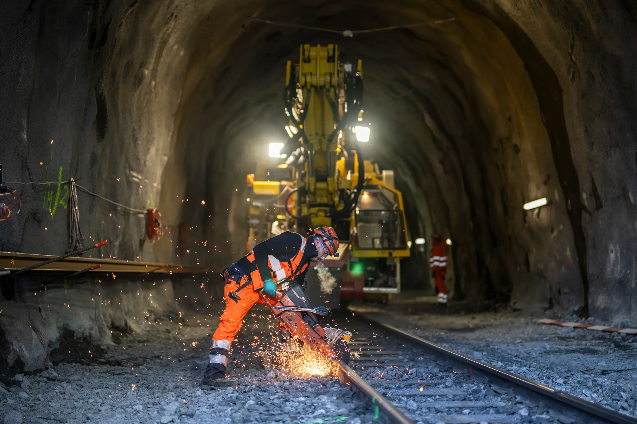Arbeiter in oranger Schutzkleidung schneidet mit einem Trennschleifer Gleise in einem Tunnel. Im Hintergrund eine Baumaschine