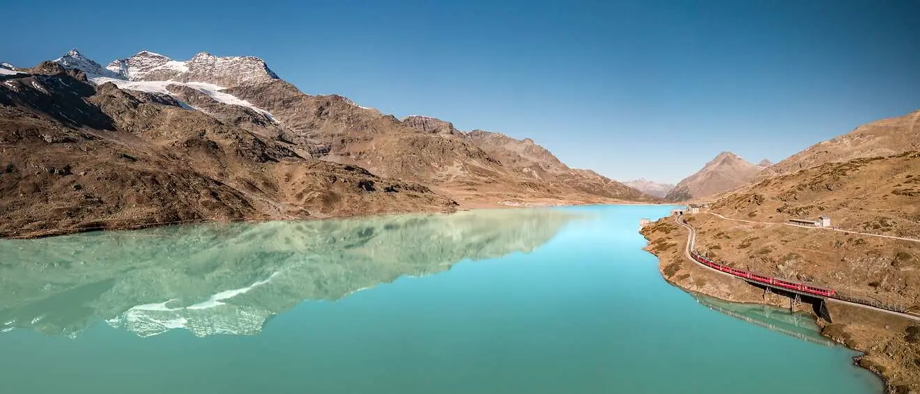 Ein roter Zug fährt am türkisblauen Lago Bianco entlang, im Hintergrund Berge und Gletscher, die sich im See spiegeln.