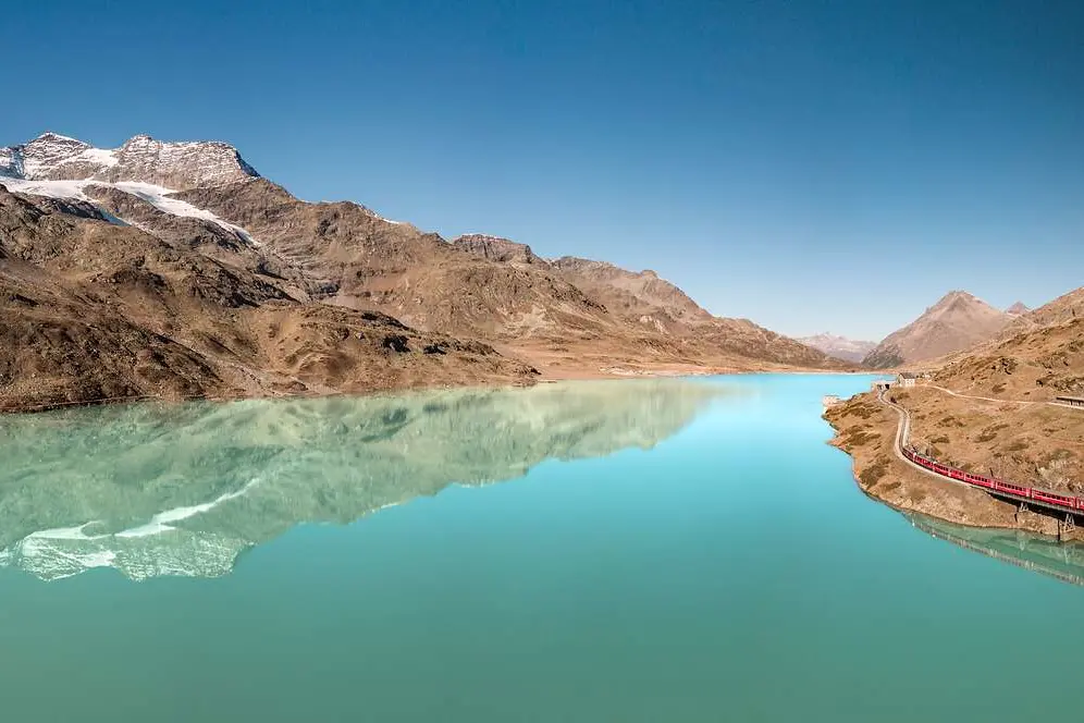 Ein roter Zug fährt am türkisblauen Lago Bianco entlang, im Hintergrund Berge und Gletscher, die sich im See spiegeln.