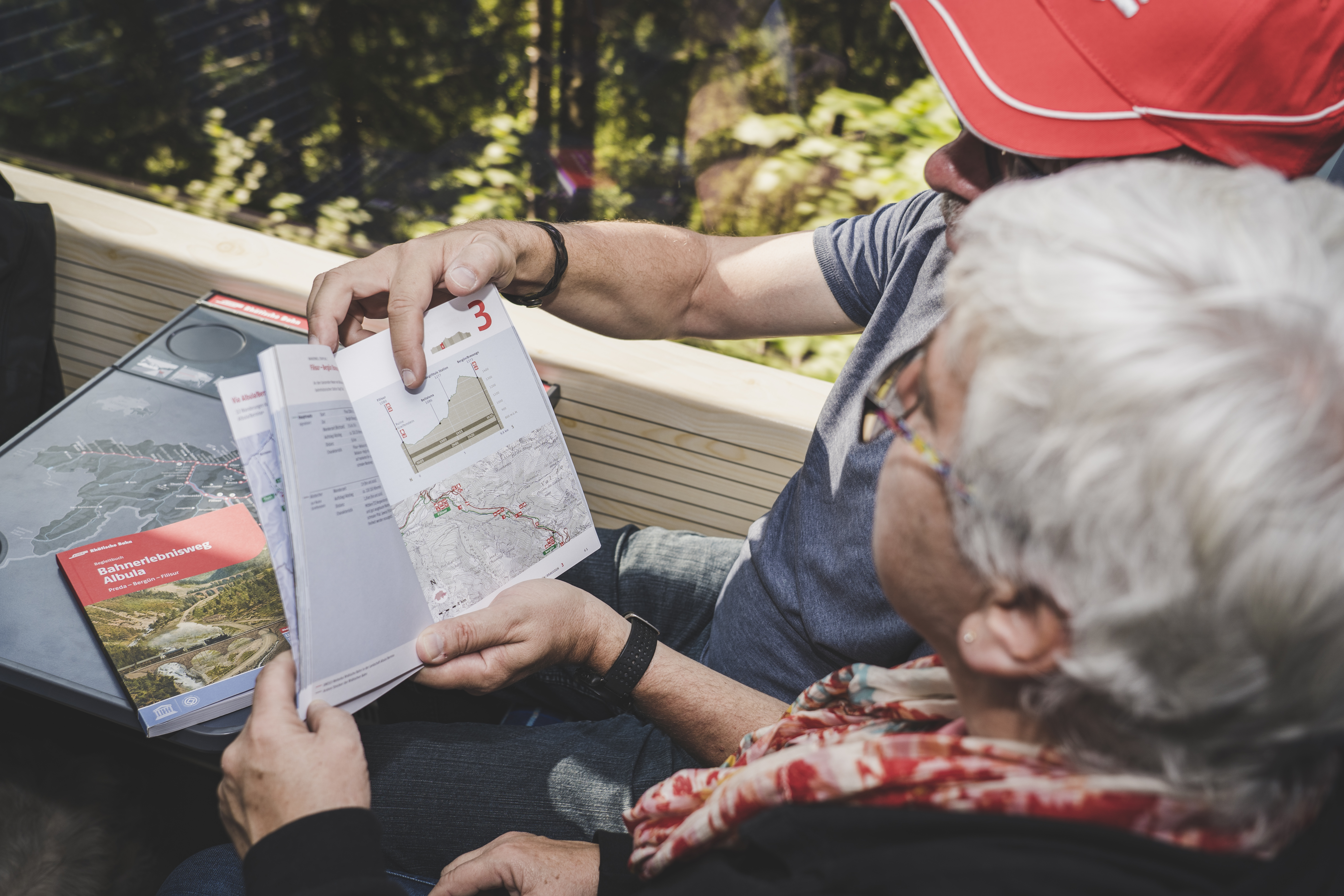 Zwei Personen sitzen im RhB-Zug und schauen sich das Begleitbuch vom Bahnerlebnisweg Albula an.
