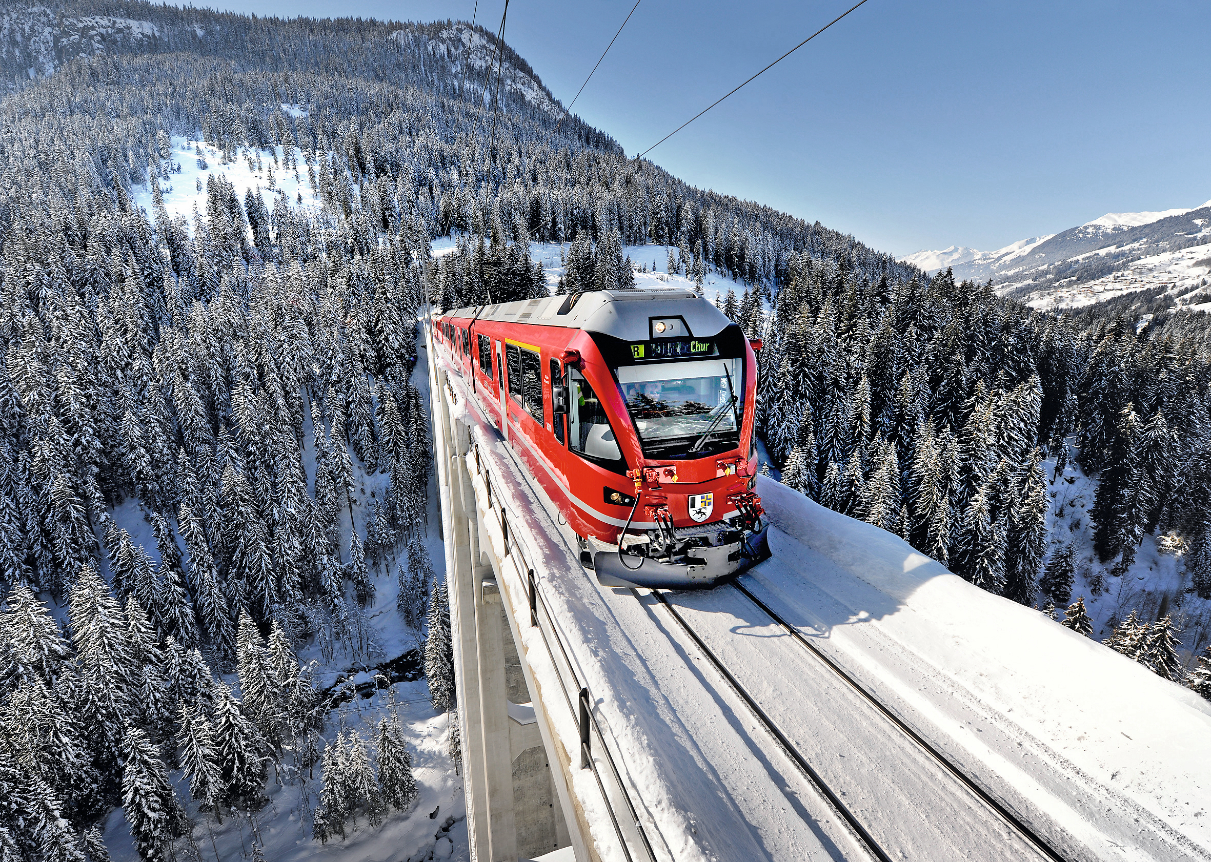 Ein Regionalzug der Rhätischen Bahn fährt auf dem Langwieserviadukt, im Hintergrund schneebedeckte Berge und Wälder.