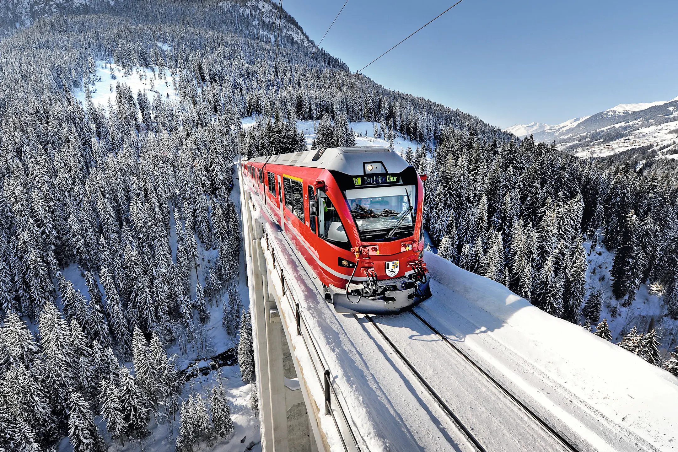 Ein Regionalzug der Rhätischen Bahn fährt auf dem Langwieserviadukt, im Hintergrund schneebedeckte Berge und Wälder.