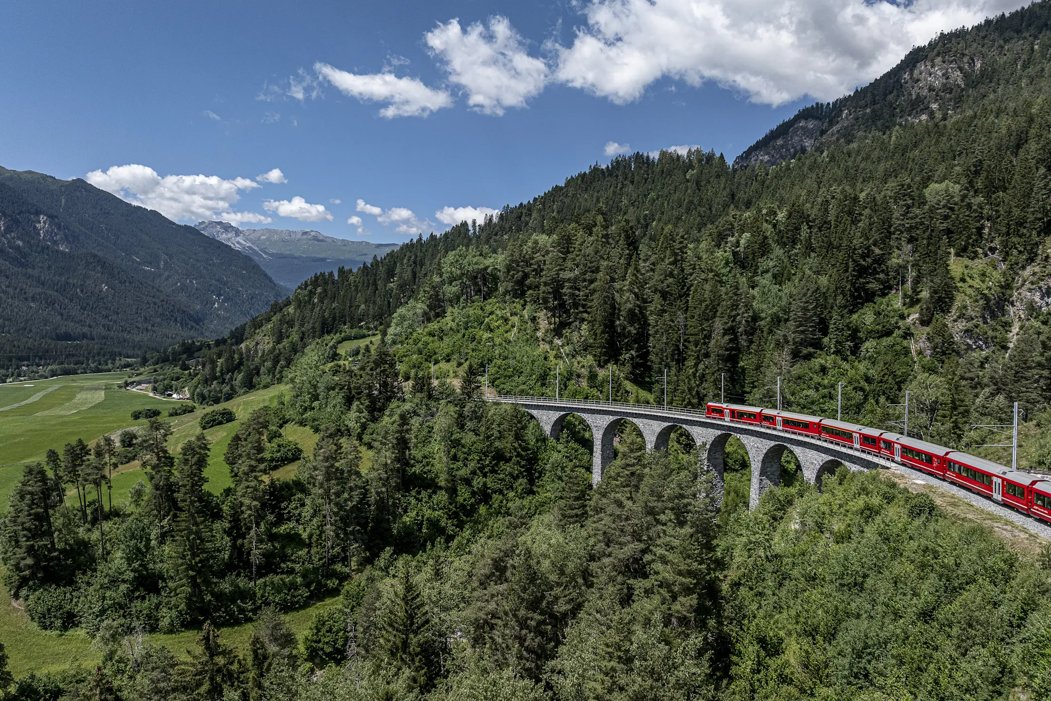 Ein roter Zug überquert den Schmittnertobelviadukt in einer grünen, bergigen Landschaft unter blauem Himmel.