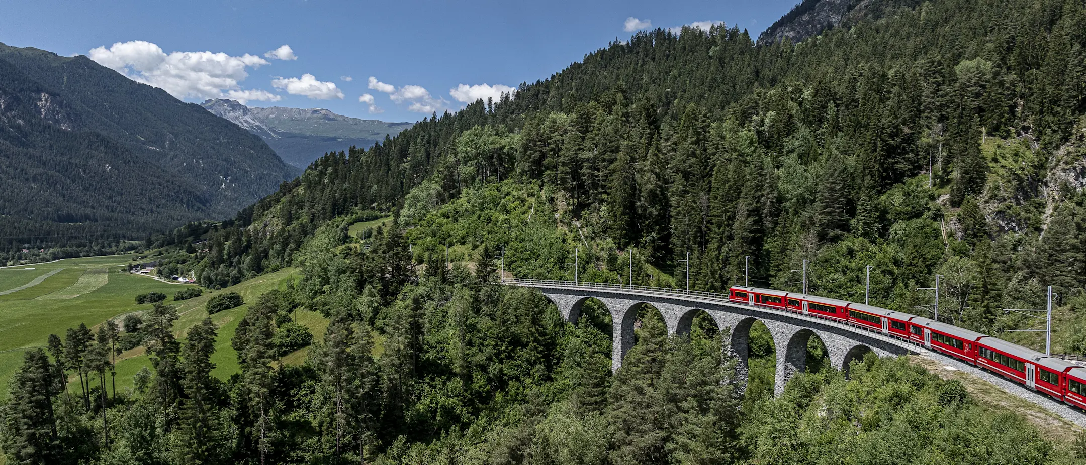 Ein roter Zug überquert den Schmittnertobelviadukt in einer grünen, bergigen Landschaft unter blauem Himmel.