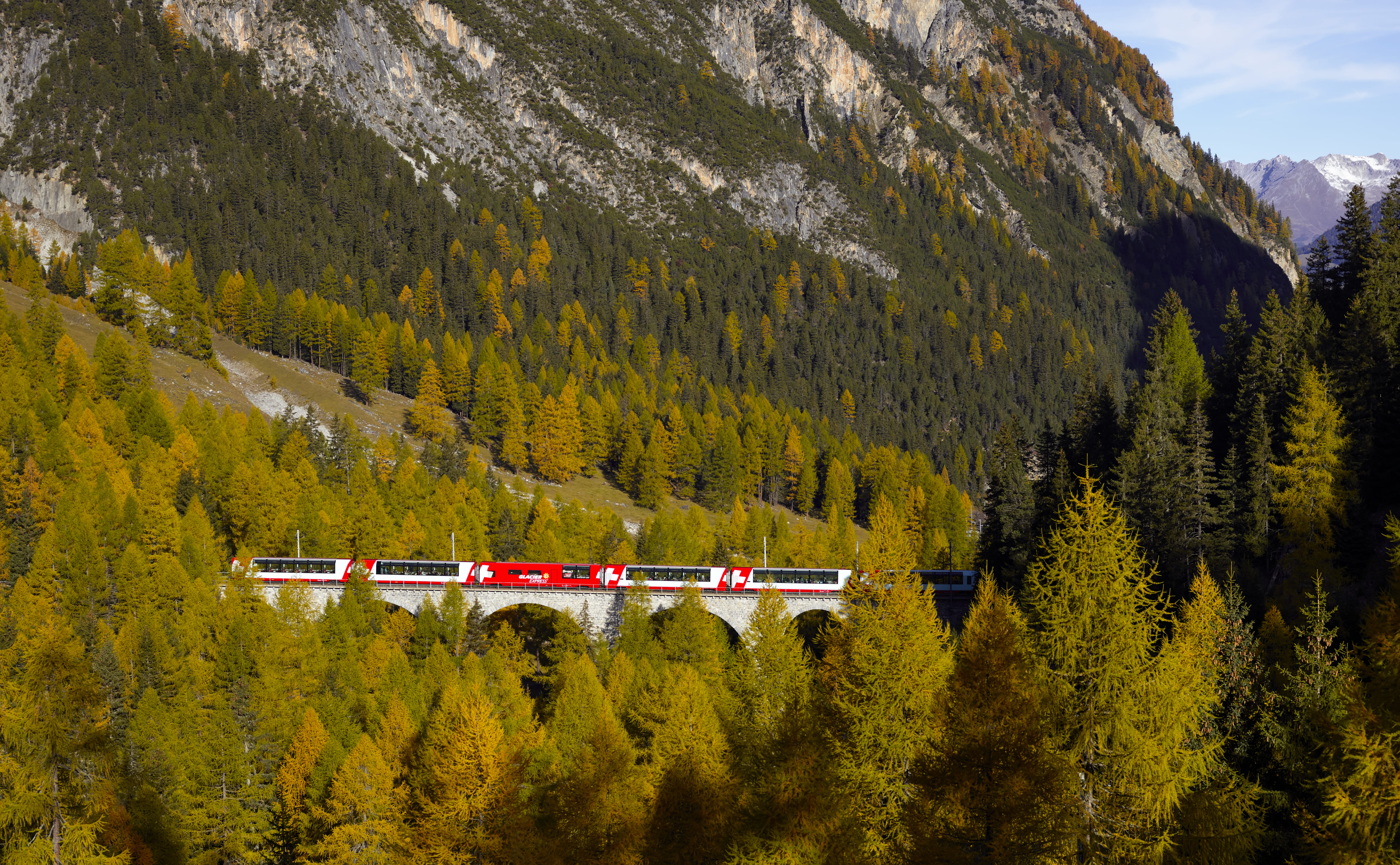 The Glacier Express travels over a viaduct through autumnal forests in the Albula Valley, surrounded by high mountain slopes.