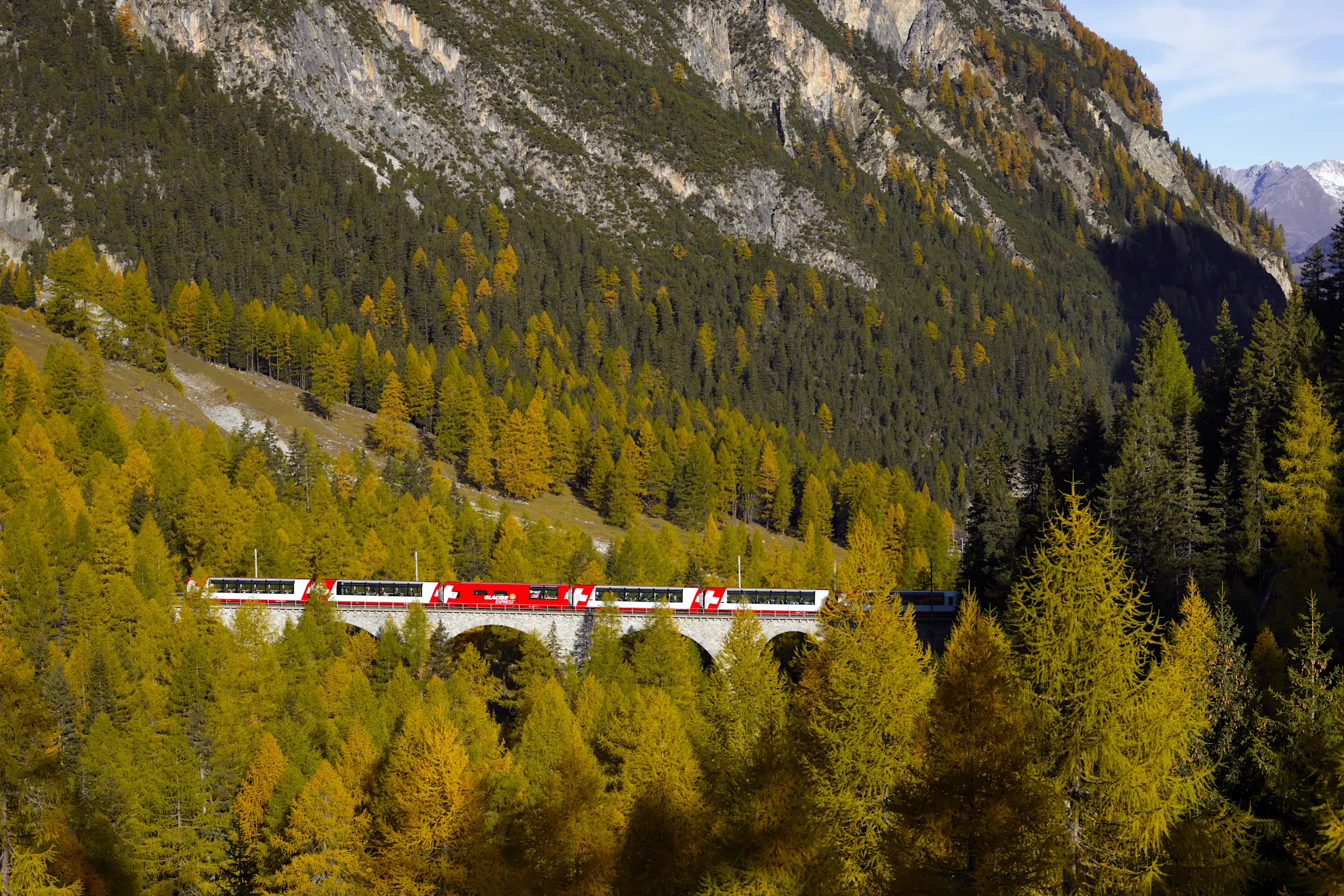 Der Glacier Express fährt über ein Viadukt durch herbstlich gefärbte Wälder im Albulatal, umgeben von hohen Bergflanken.