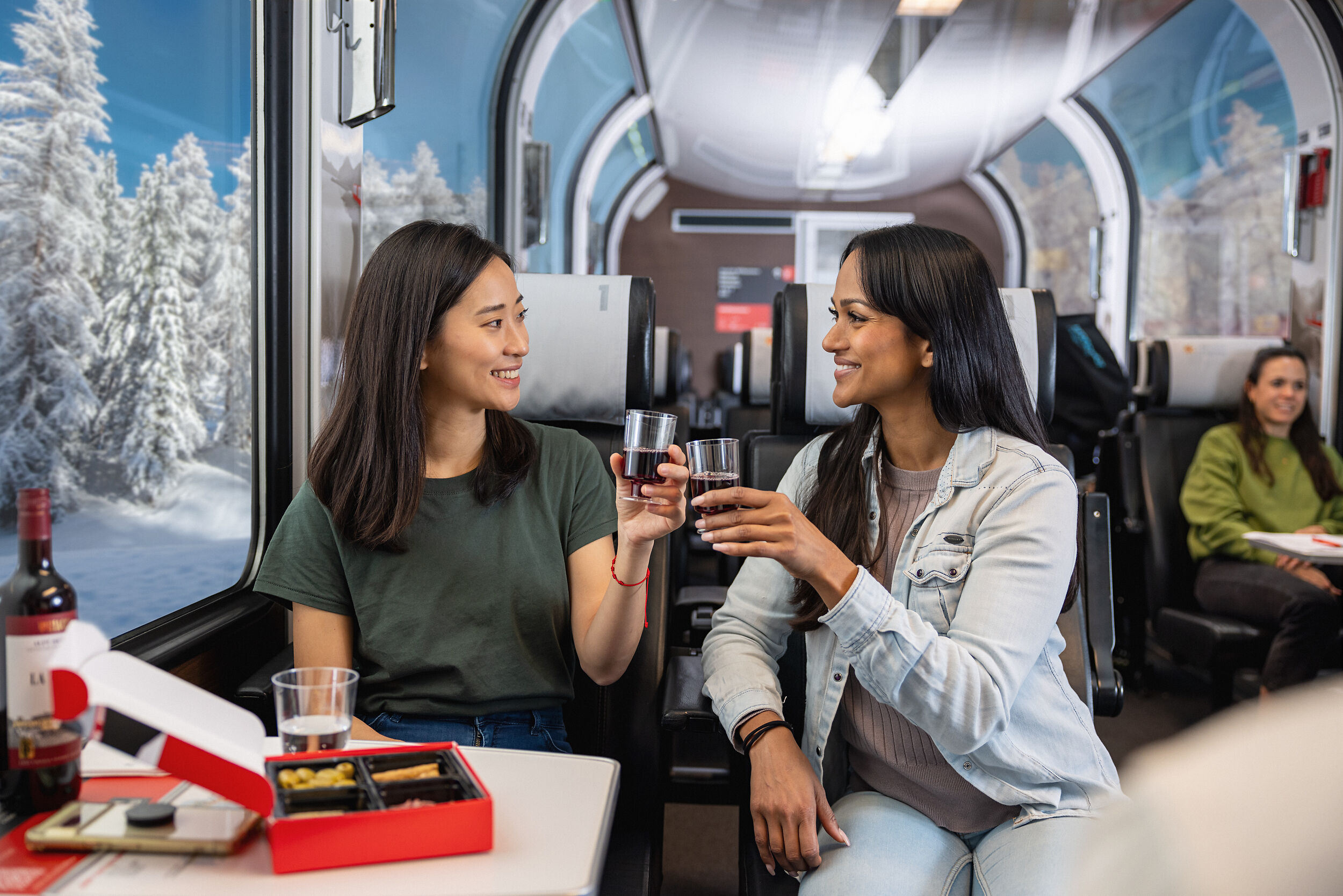 Zwei Frauen stossen im Bernina Express mit einem Glas Rotwein an. Im Hintergrund sieht man die winterliche Landschaft. 