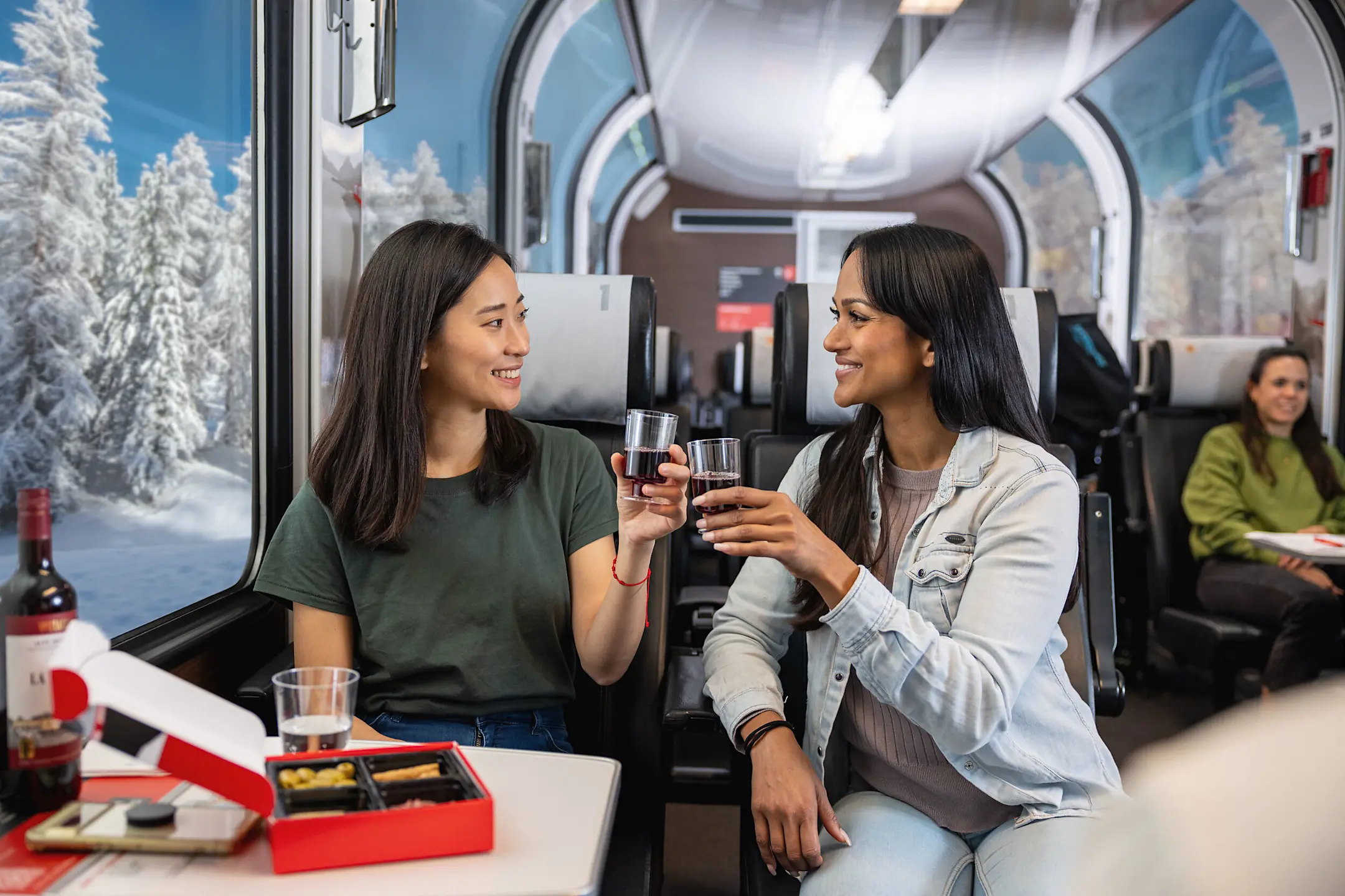 Zwei Frauen stossen im Bernina Express mit einem Glas Rotwein an. Im Hintergrund sieht man die winterliche Landschaft.