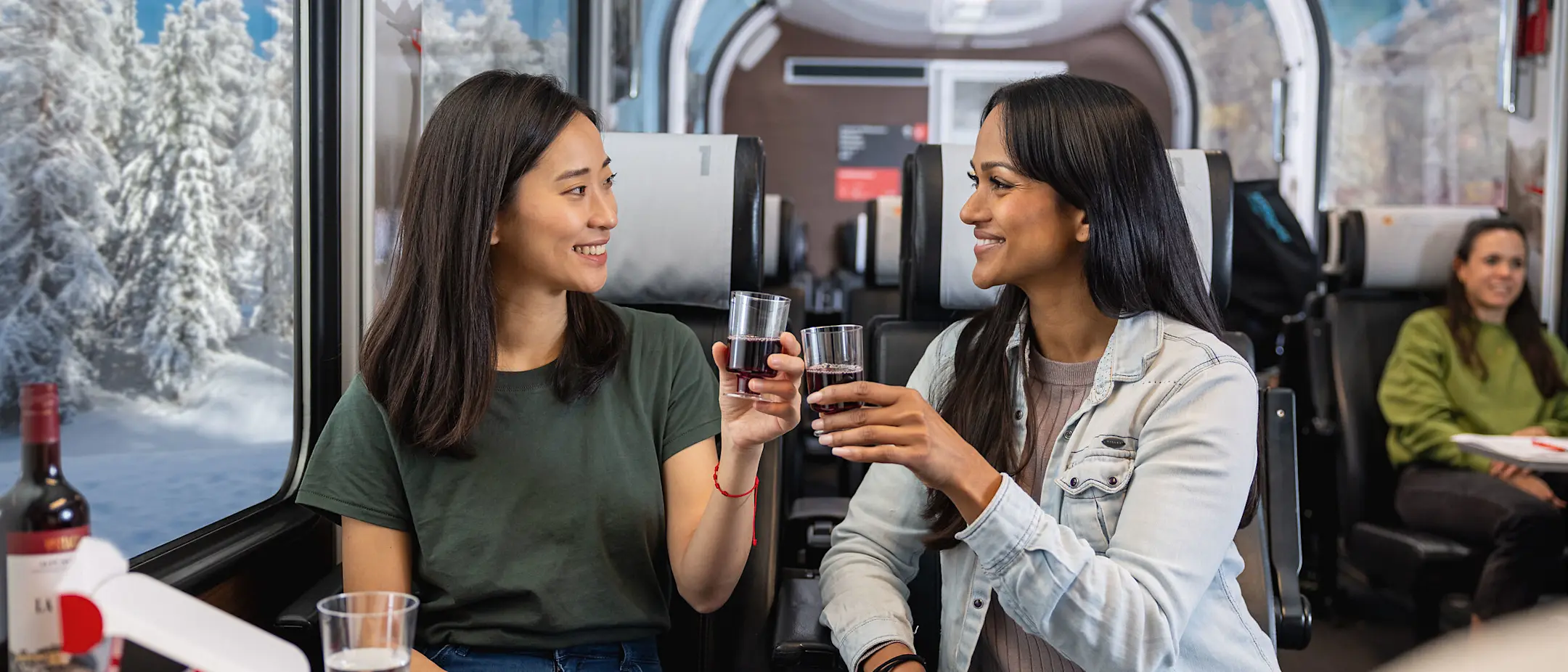 Zwei Frauen stossen im Bernina Express mit einem Glas Rotwein an. Im Hintergrund sieht man die winterliche Landschaft.