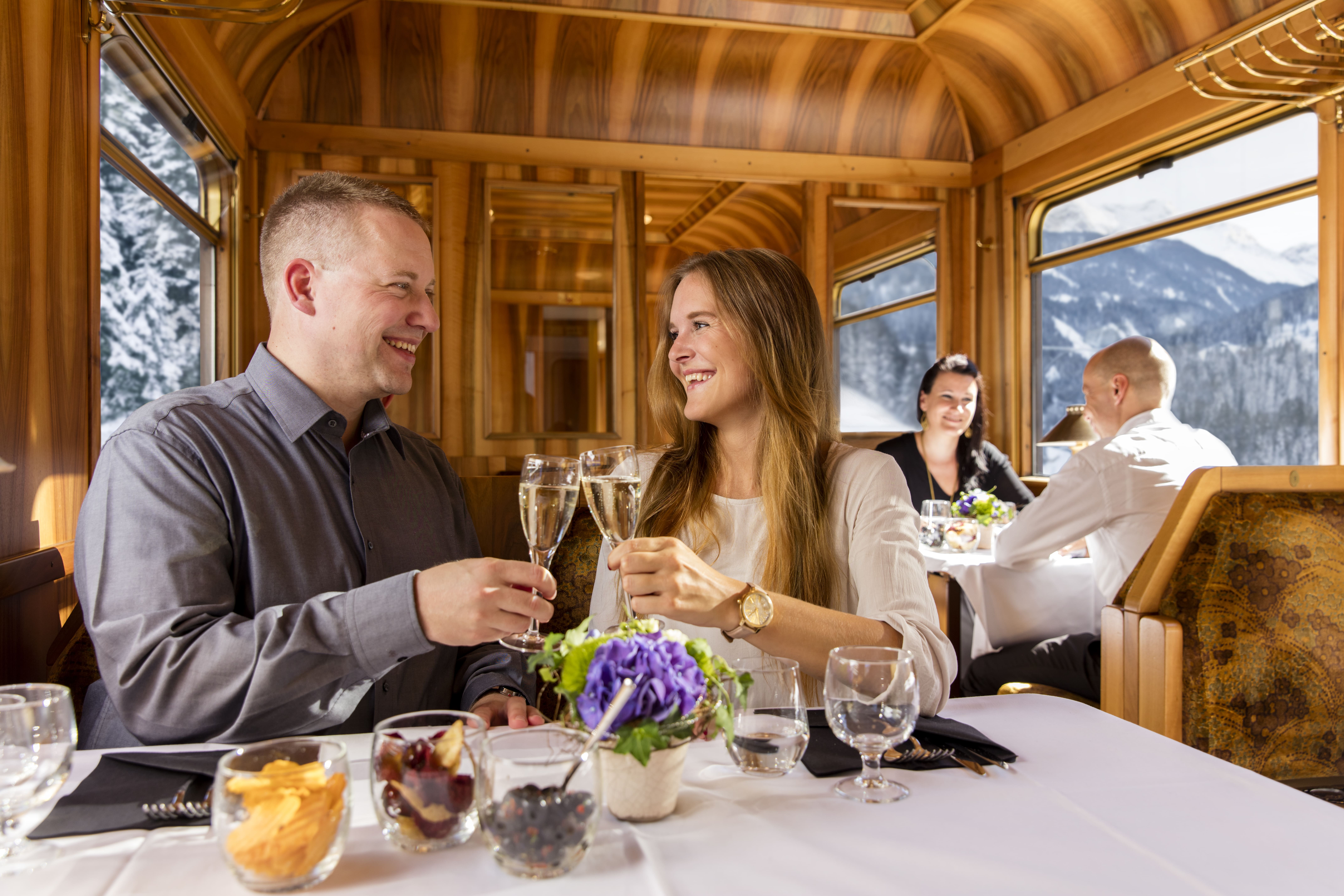 Ein Paar sitzt im Speisewagen Gourmino, stößt mit Sektgläsern an. Im Hintergrund genießt ein weiteres Paar das Essen.